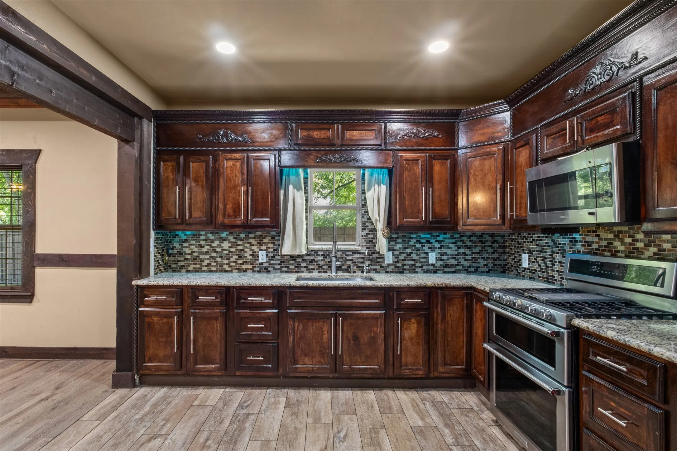 Kitchen featuring wood-finish flooring, custom dark wood cabinetry with ornate detailing, a mosaic tile backsplash, and stone countertops