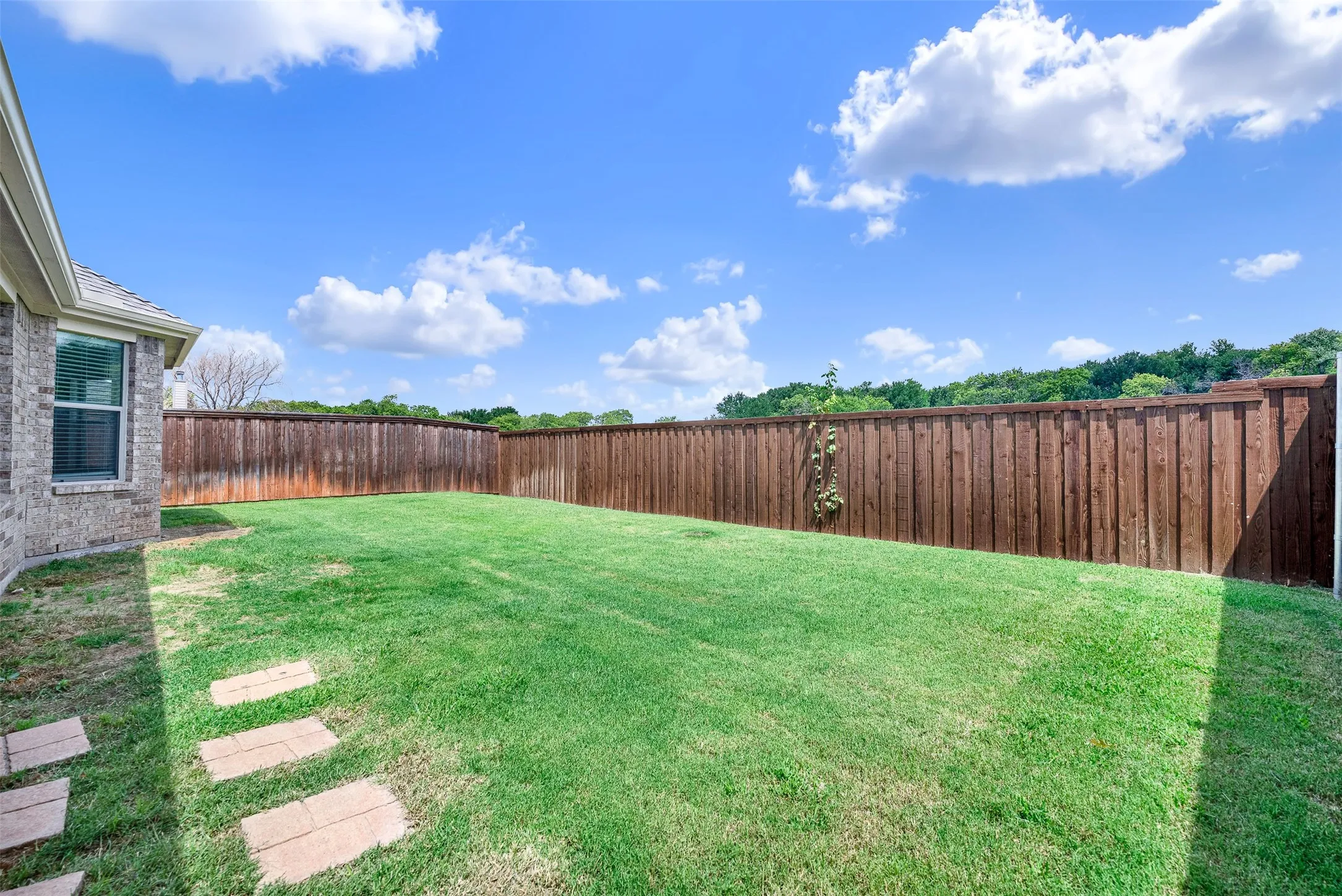 Expansive green lawn surrounded by a privacy fence, featuring a brick exterior wall and stone stepping pavers