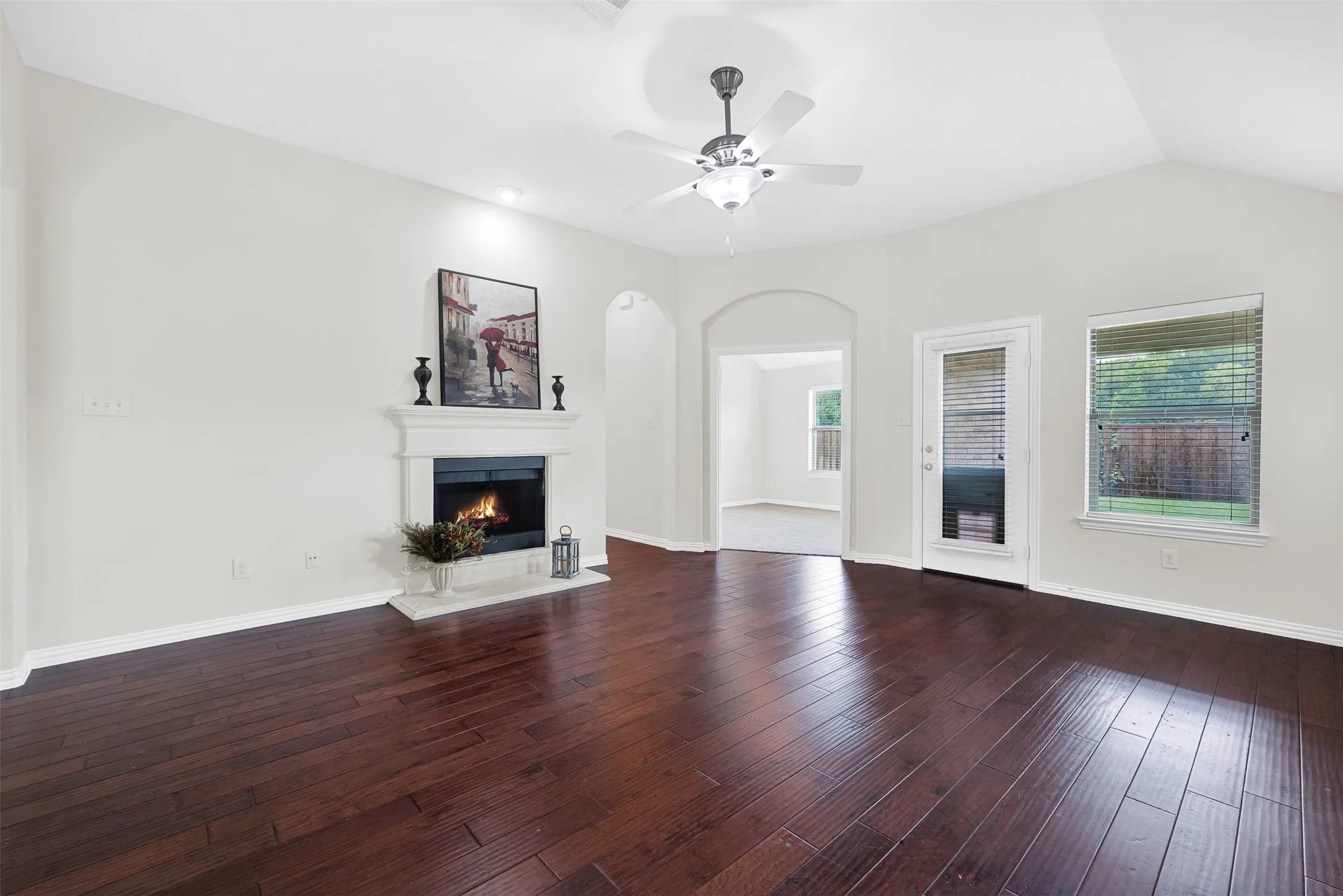 Living area featuring rich wood-finish flooring, a decorative fireplace with a mantel, an arched entry, a glass-paned exterior door, and a ceiling fan with integrated lighting