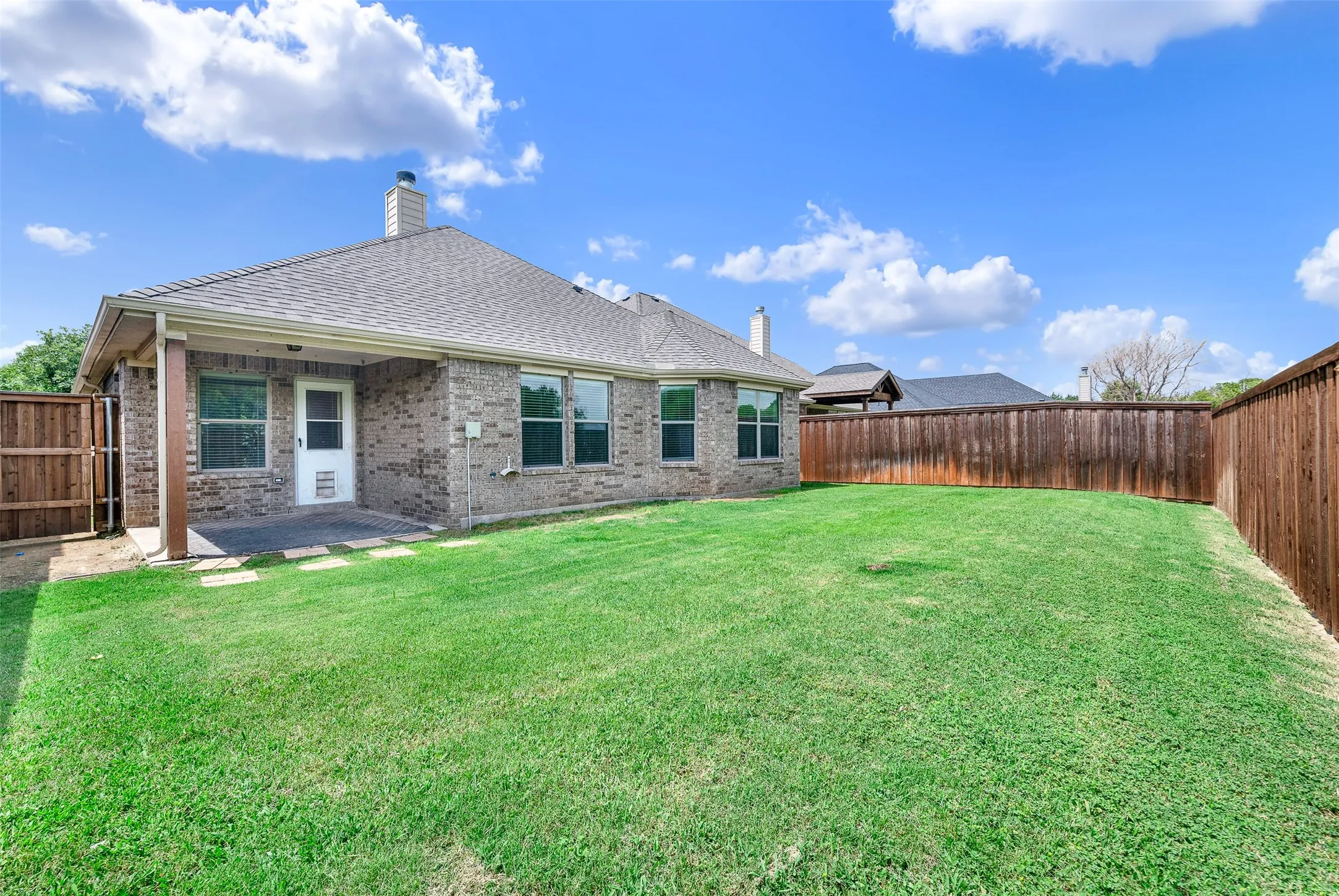 Expansive fenced backyard featuring a covered patio with tile flooring, a brick exterior, multiple windows, and a shingled roof