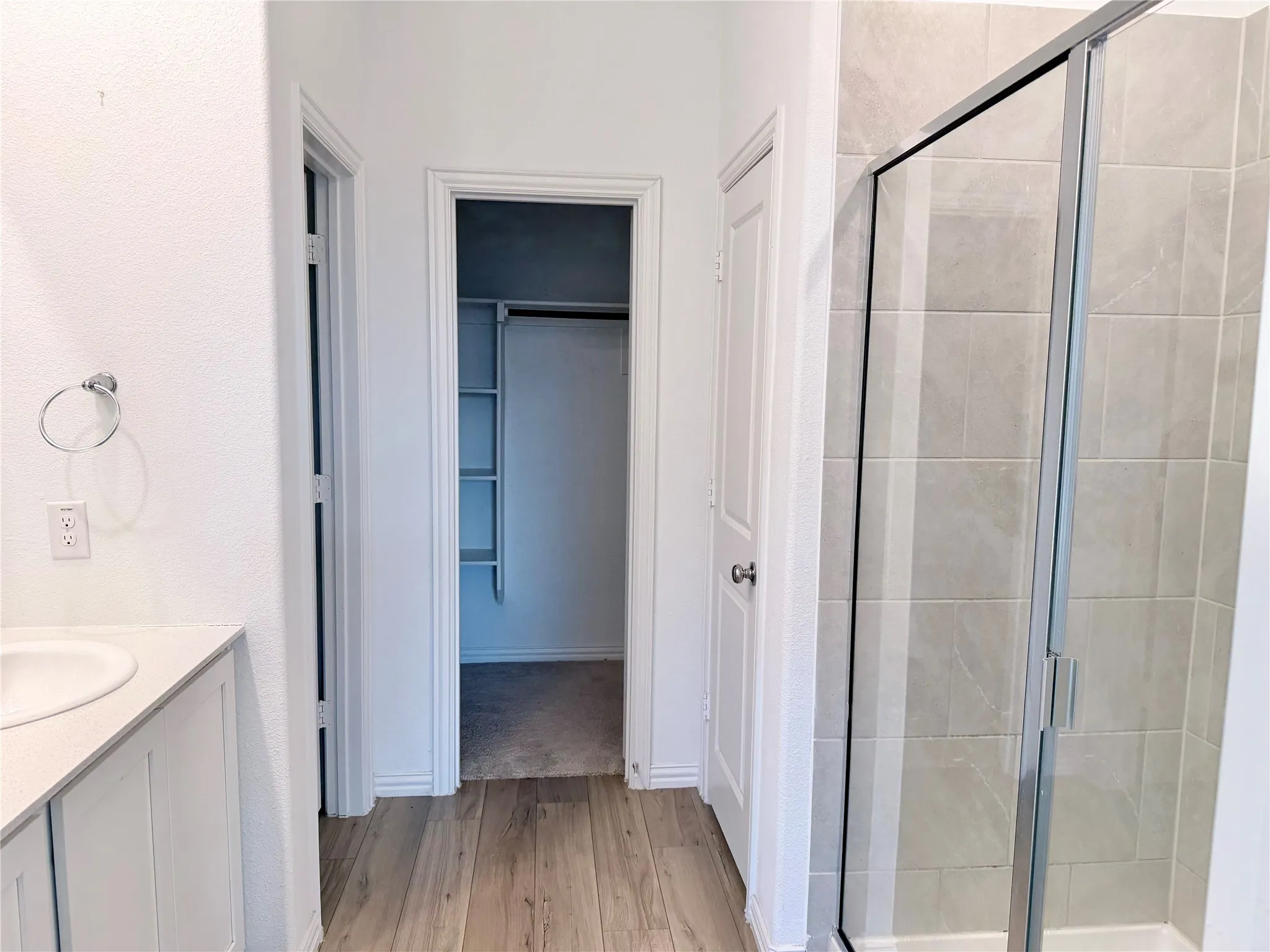 Bathroom featuring a glass-enclosed shower with large format tile surround, wood-finish flooring, a white vanity with an undermount sink and light-toned countertop, and an adjacent walk-in closet with built-in shelving