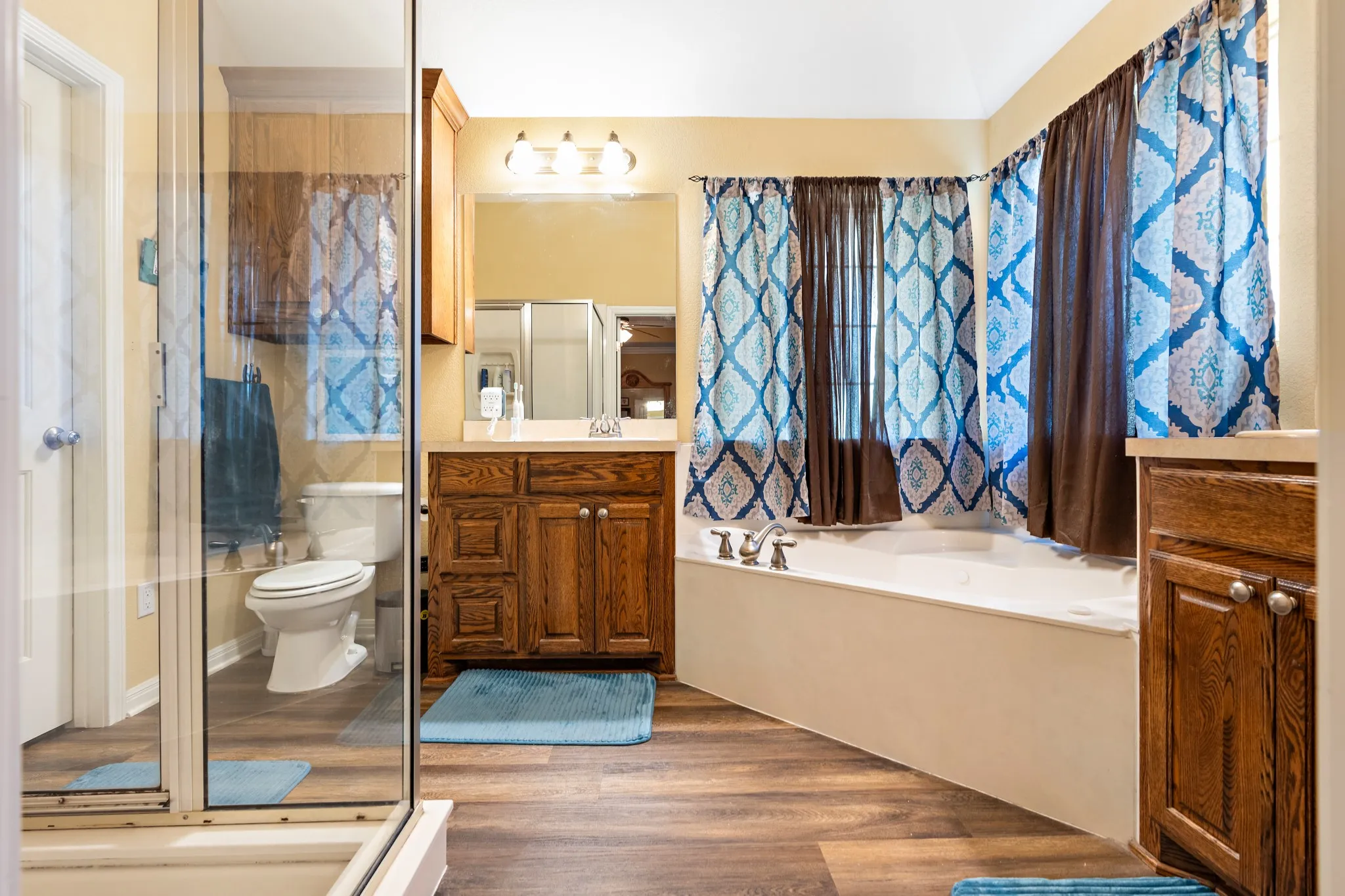 Bathroom featuring a corner soaking tub, a separate glass-enclosed shower, wood-finish flooring, a vanity with a light-toned countertop, and an additional built-in storage cabinet
