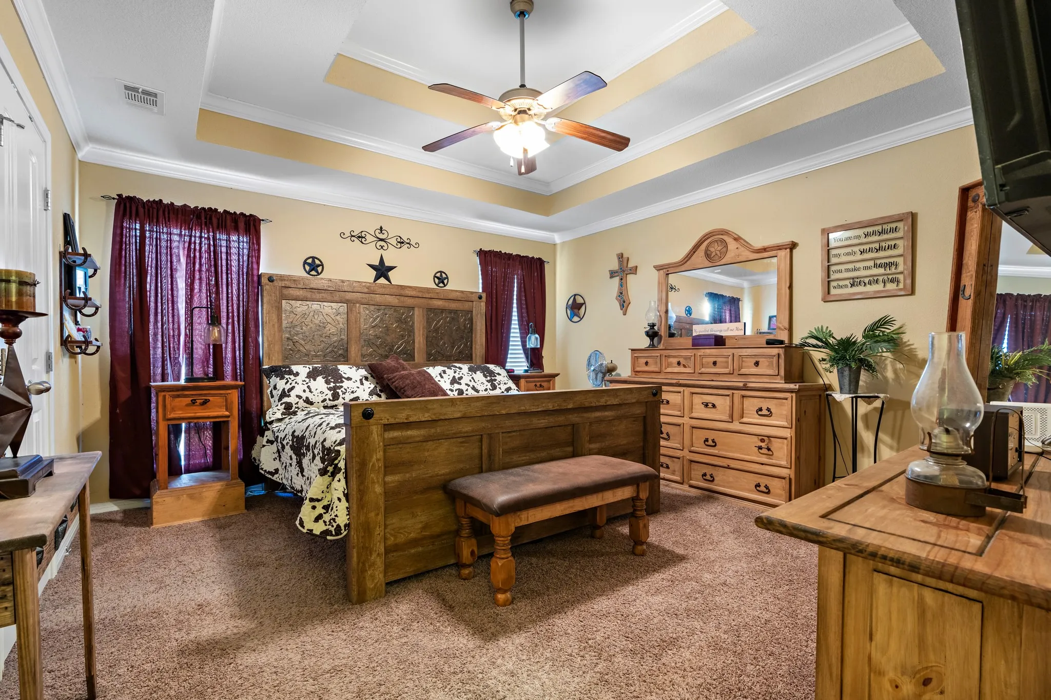 Spacious bedroom featuring a tray ceiling with a ceiling fan, neutral carpet flooring, and multiple window openings with dark drapery