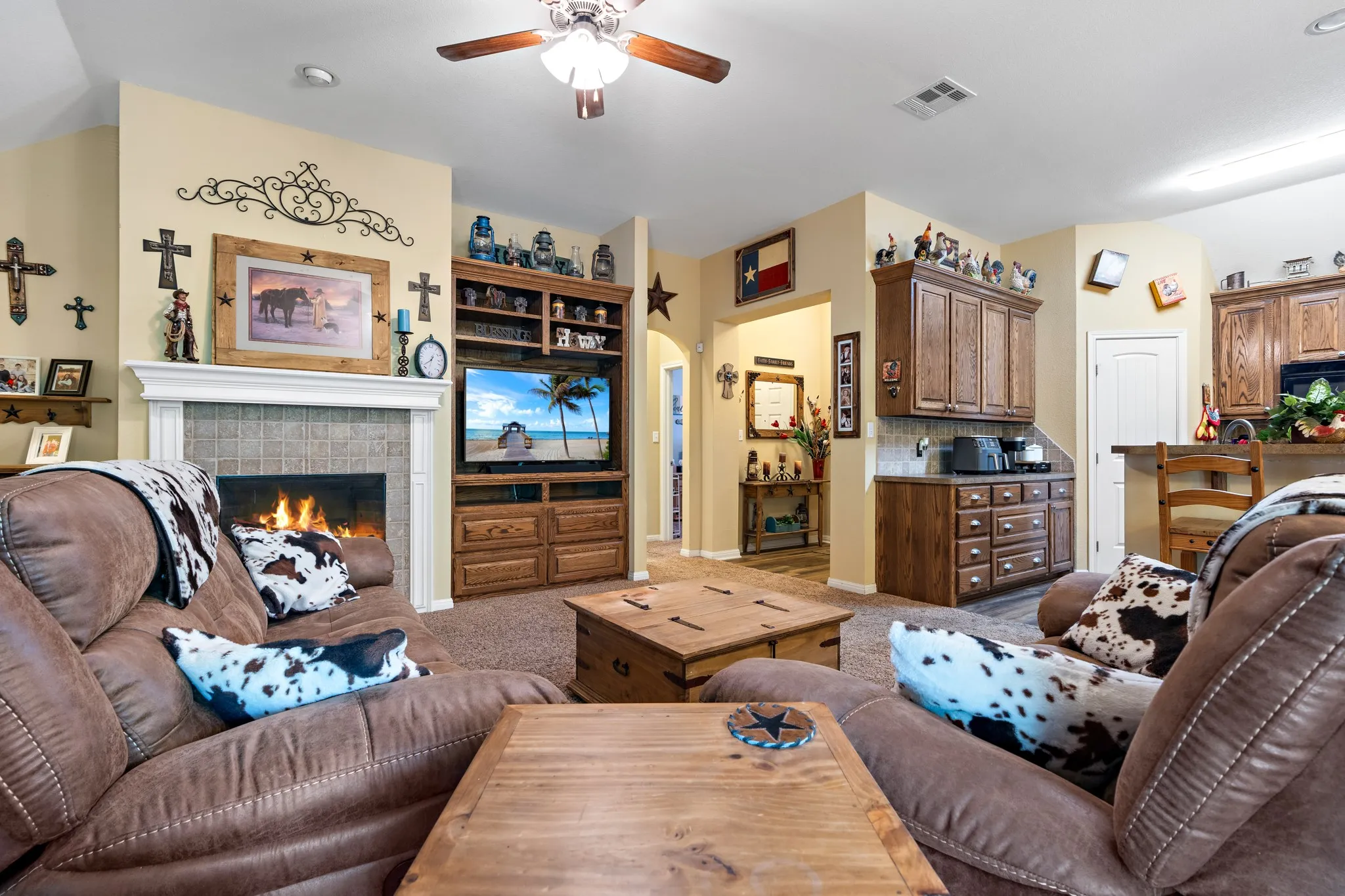 Carpeted living area featuring a tile-surround fireplace with a white mantel, built-in entertainment center with shelving, ceiling fan, and an open layout to a kitchen area with wood-finish cabinetry