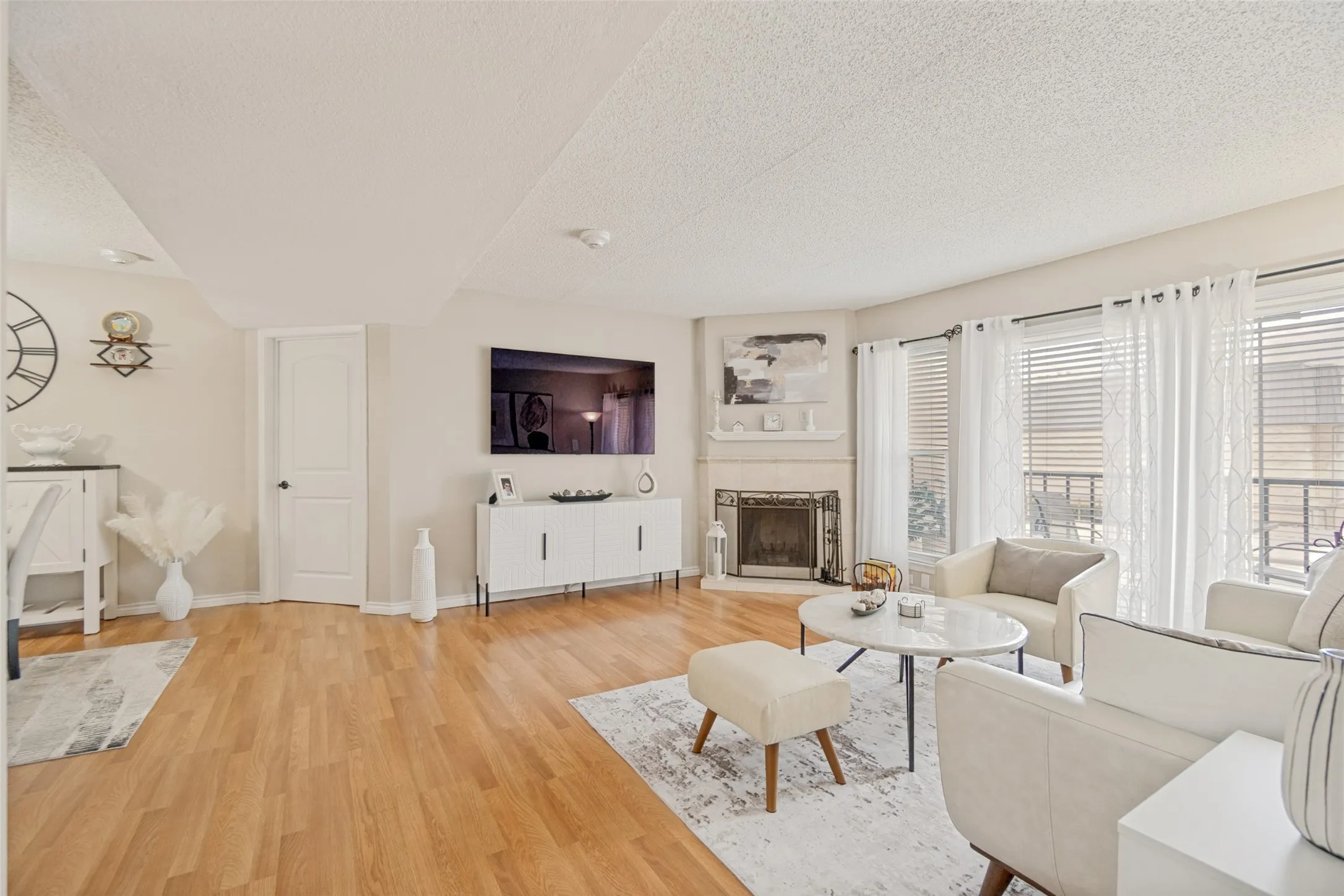 Living area with wood-finish flooring, a corner fireplace featuring a stone surround and mantel, and multiple windows with blinds and sheer curtains
