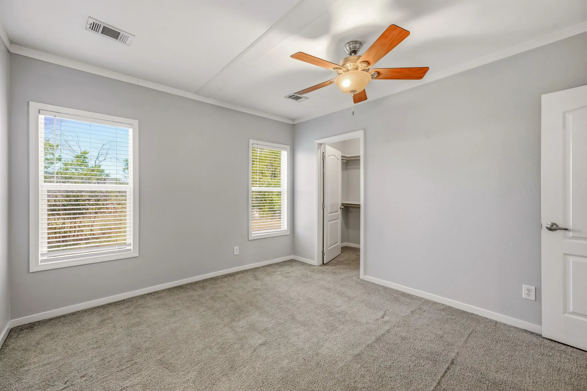 Carpeted room featuring light gray walls, white trim, two windows with blinds, an open closet with shelving, and a ceiling fan with wood-finish blades