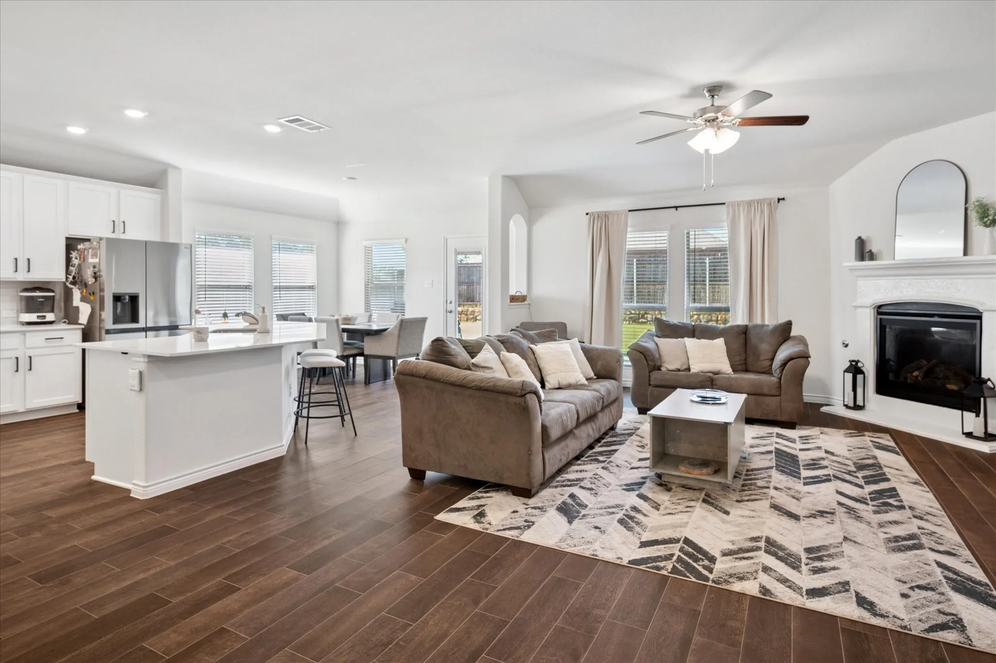 Open-concept living space featuring wood-finish flooring, a white kitchen island with seating, recessed lighting, and a fireplace with a mantel