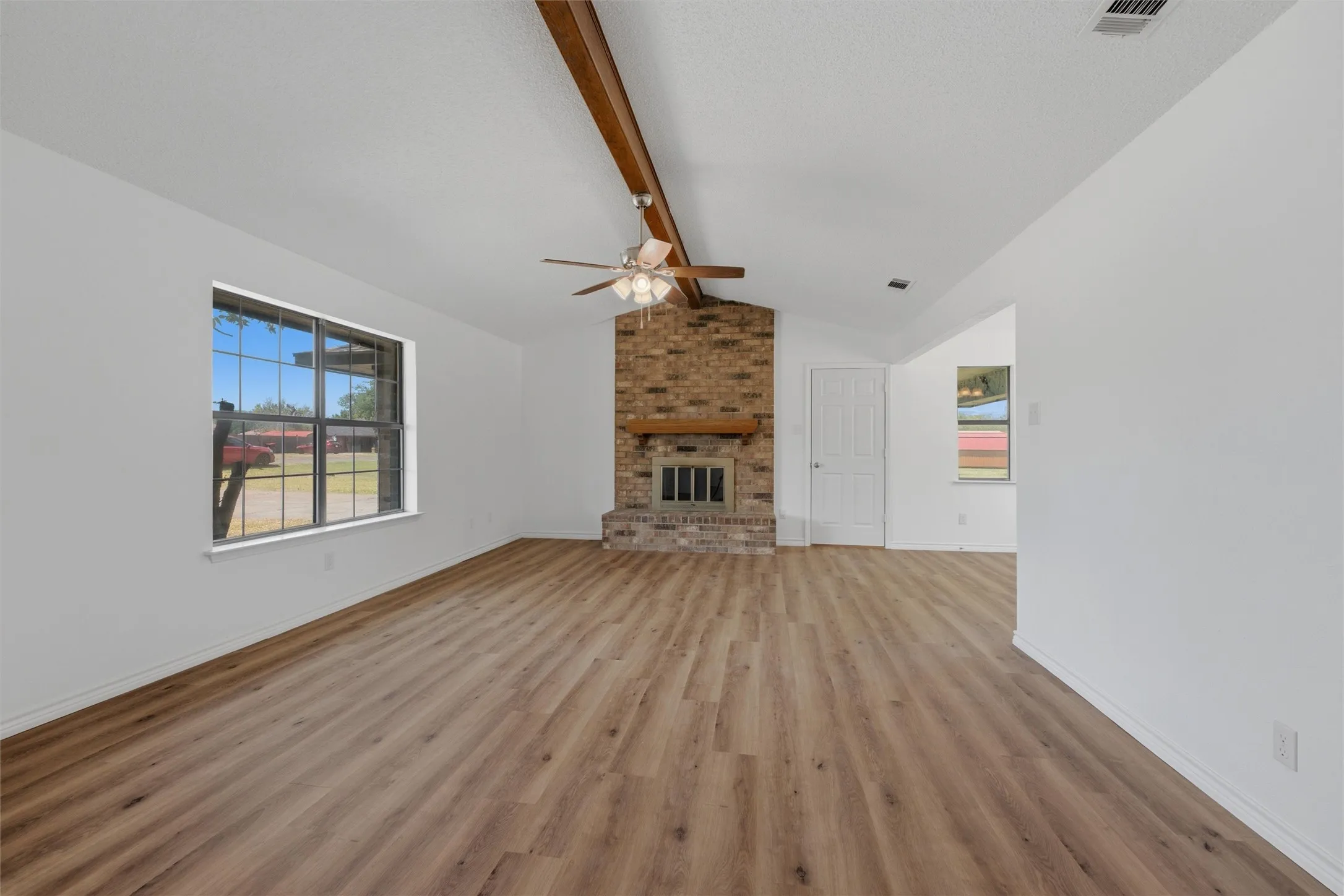 Spacious open-concept room featuring engineered wood-finish flooring, exposed wooden beam ceiling with ceiling fan, brick fireplace with chimney, large multi-pane window with natural light, and white drywall construction