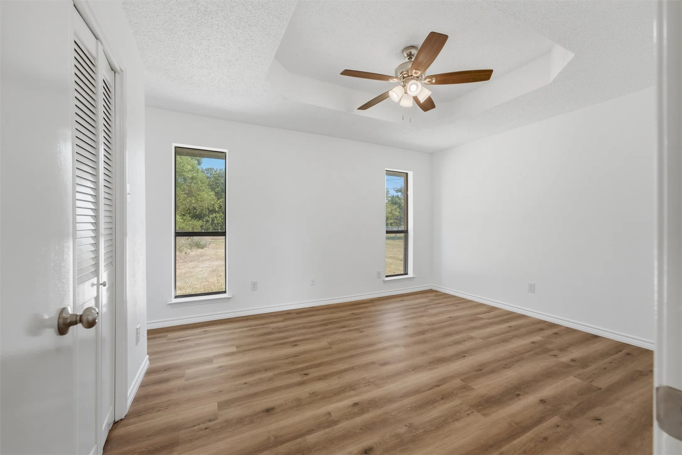 Spacious interior bedroom featuring wood-finish flooring throughout, multiple windows with natural light exposure, ceiling fan with integrated lighting, and clean white walls with fresh finishes