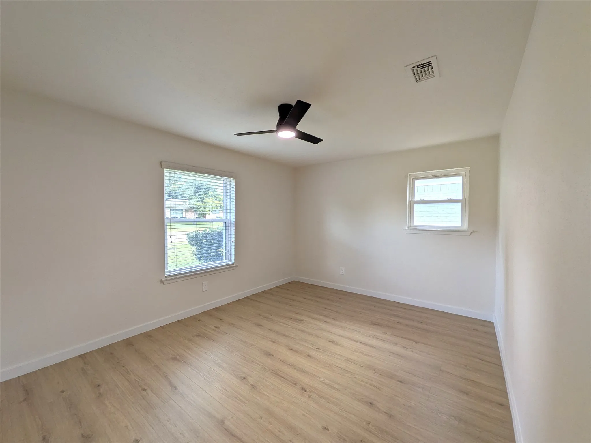 Room with wood-finish flooring, two windows with blinds, a black ceiling fan with integrated lighting, and white baseboards
