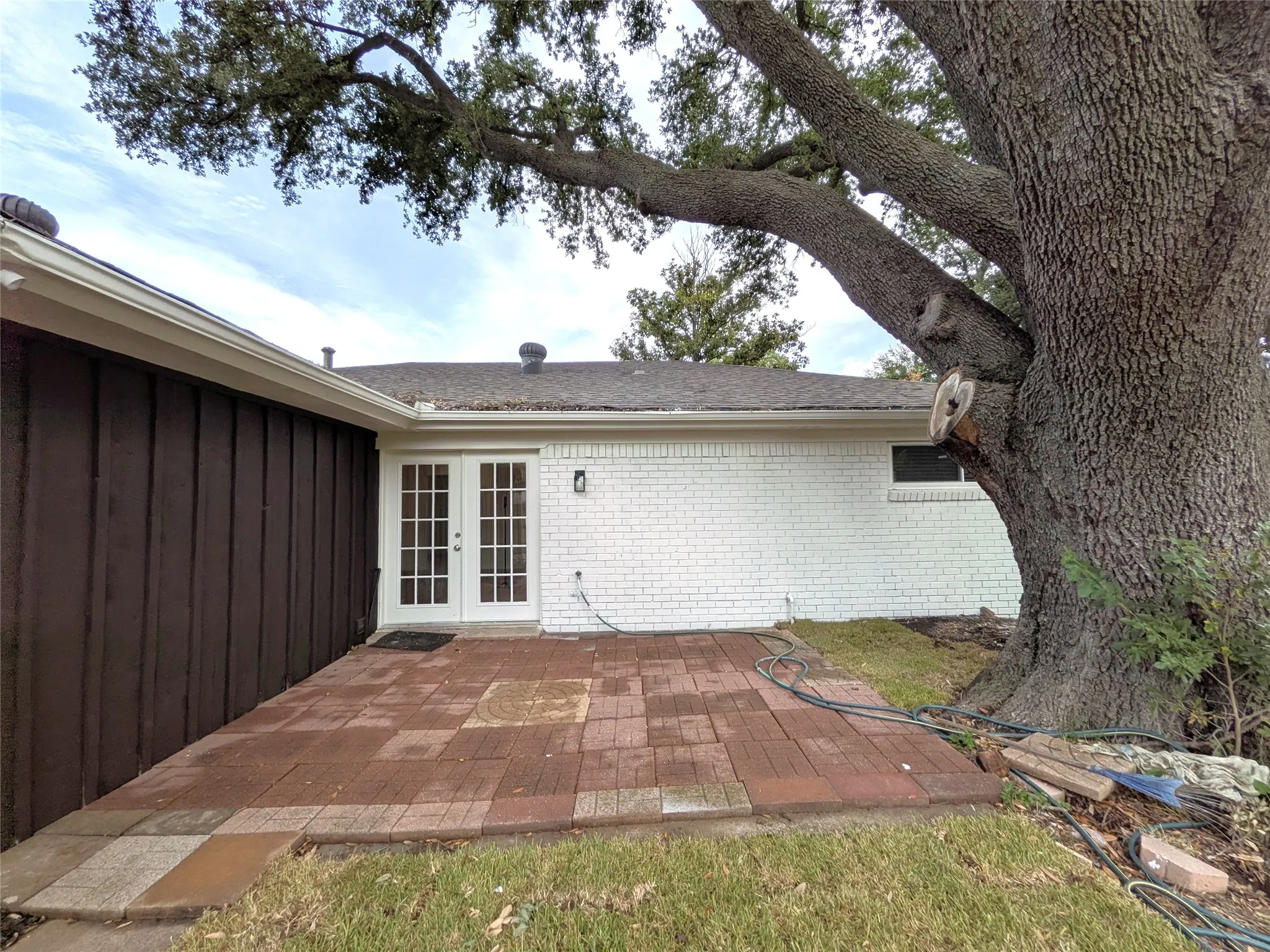 Brick paver patio with a French door entry, white painted brick facade, brown vertical siding, and an established mature tree