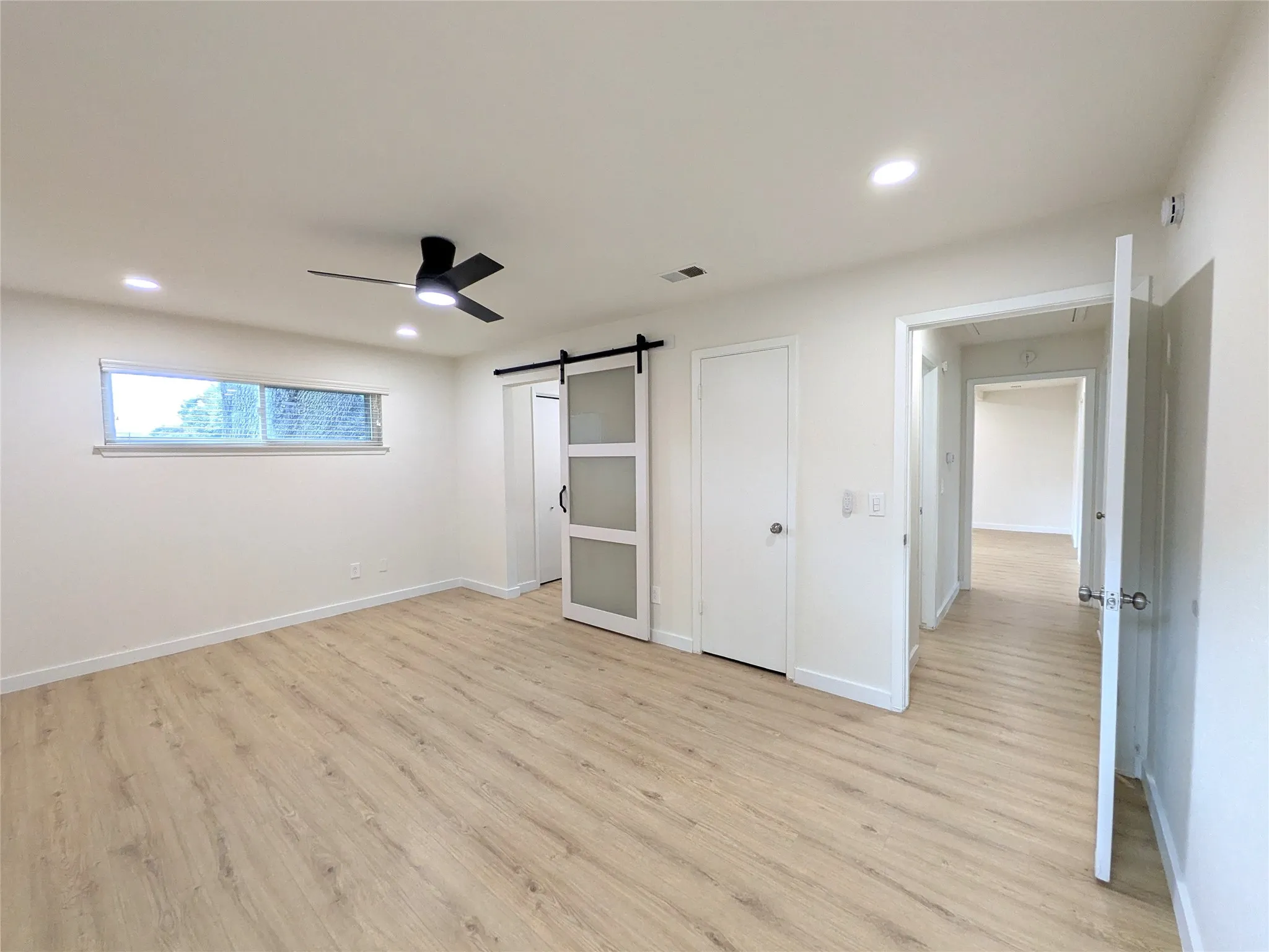 Spacious room featuring wood-finish flooring, a modern black ceiling fan, recessed lighting, and a sliding barn door with frosted glass panels