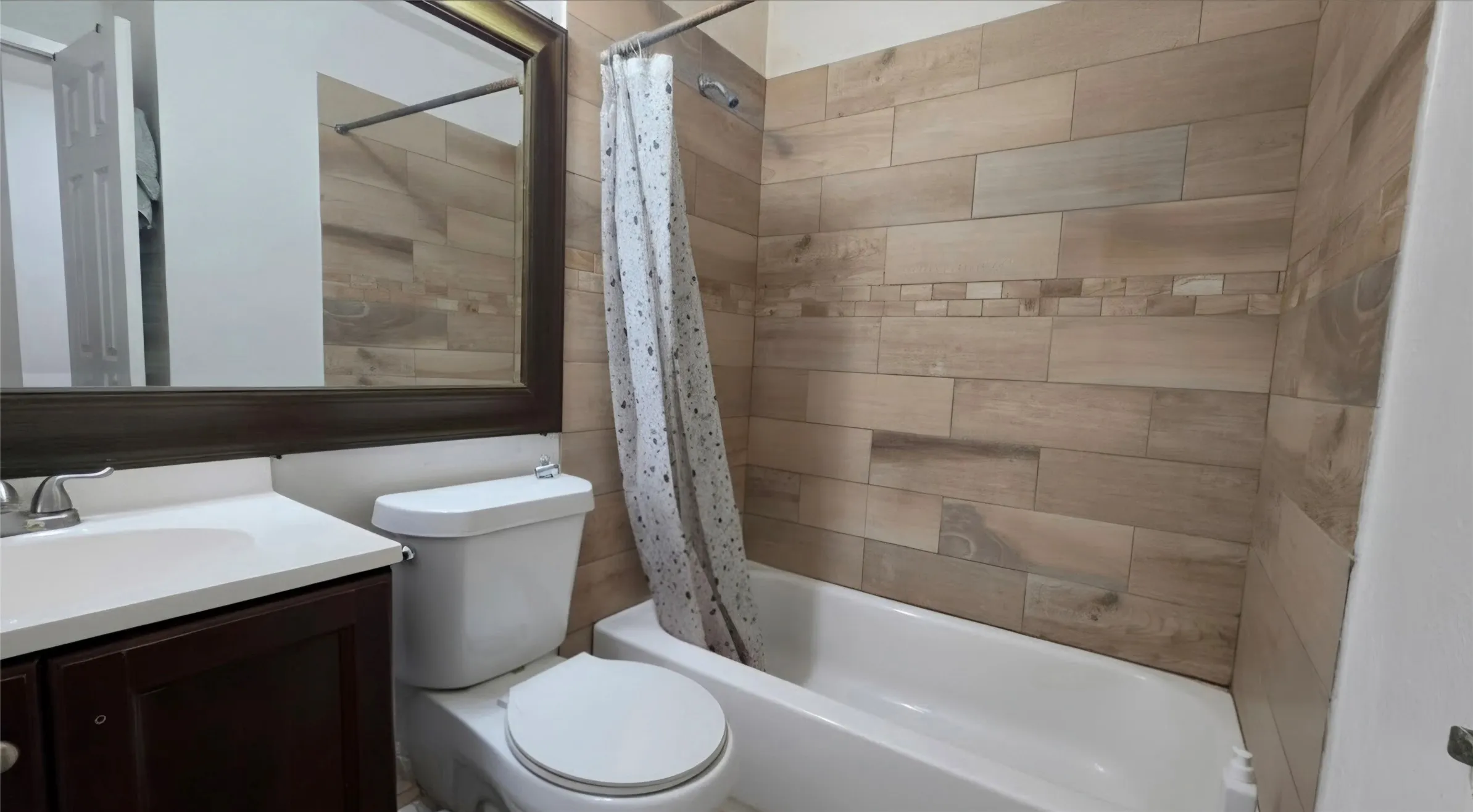Bathroom featuring a white bathtub, wood-finish tile shower surround with a decorative accent band, a white toilet, and a vanity with a white countertop and dark wood-finish cabinetry