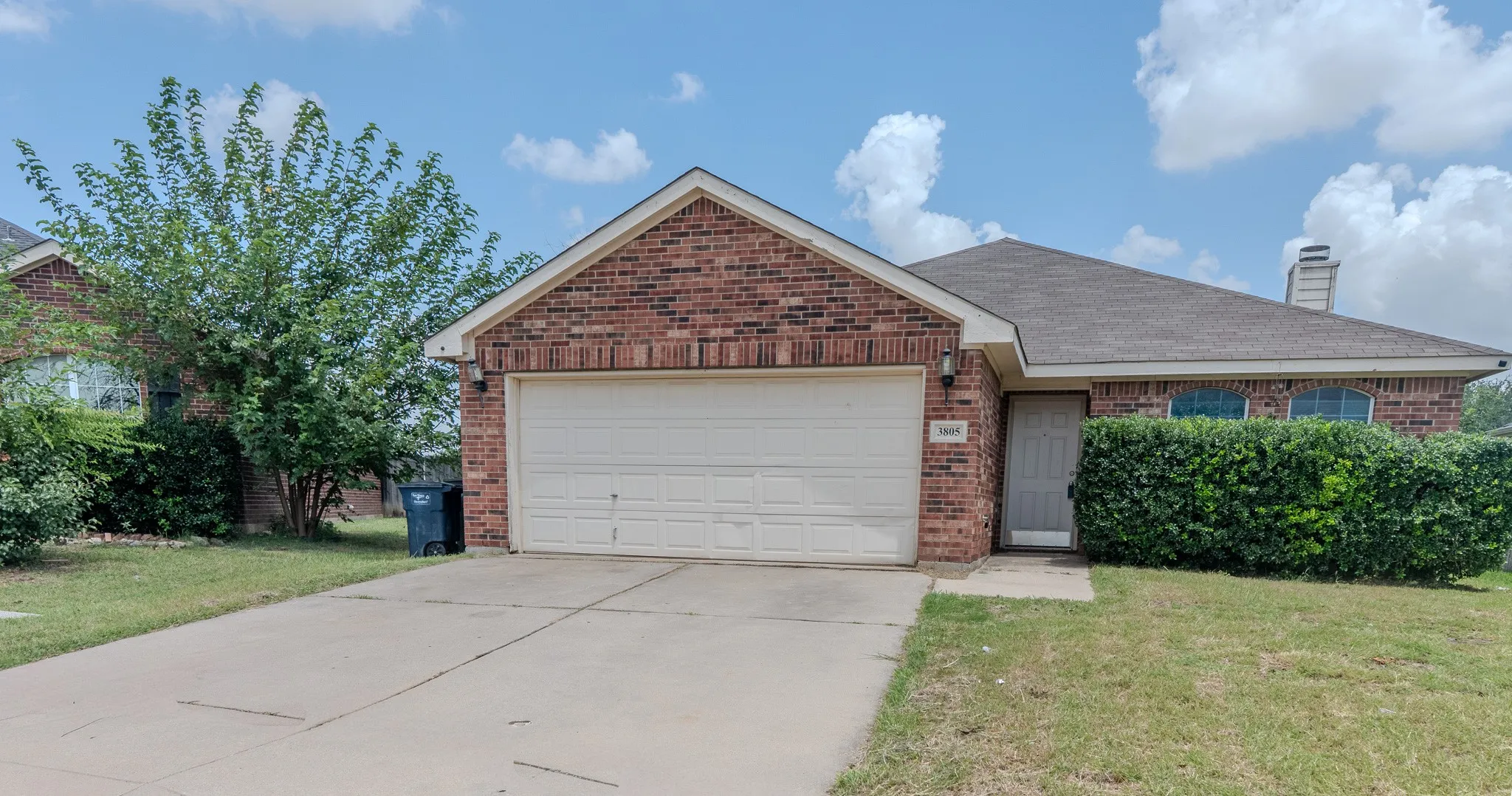 Brick facade residence featuring an attached garage, a paved driveway, and mature landscaping