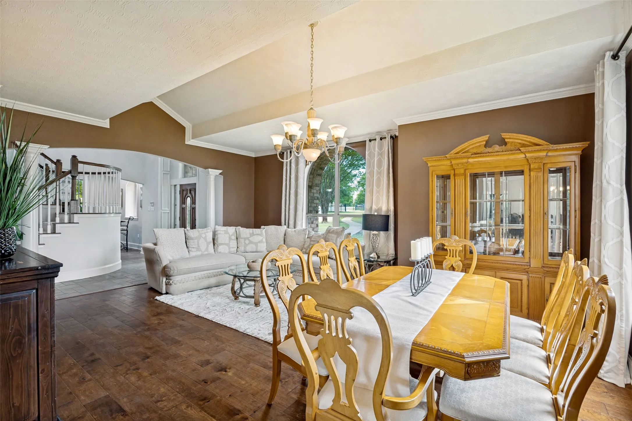Open-concept dining and living areas featuring wood-finish flooring, vaulted ceilings with crown molding, and an arched entryway