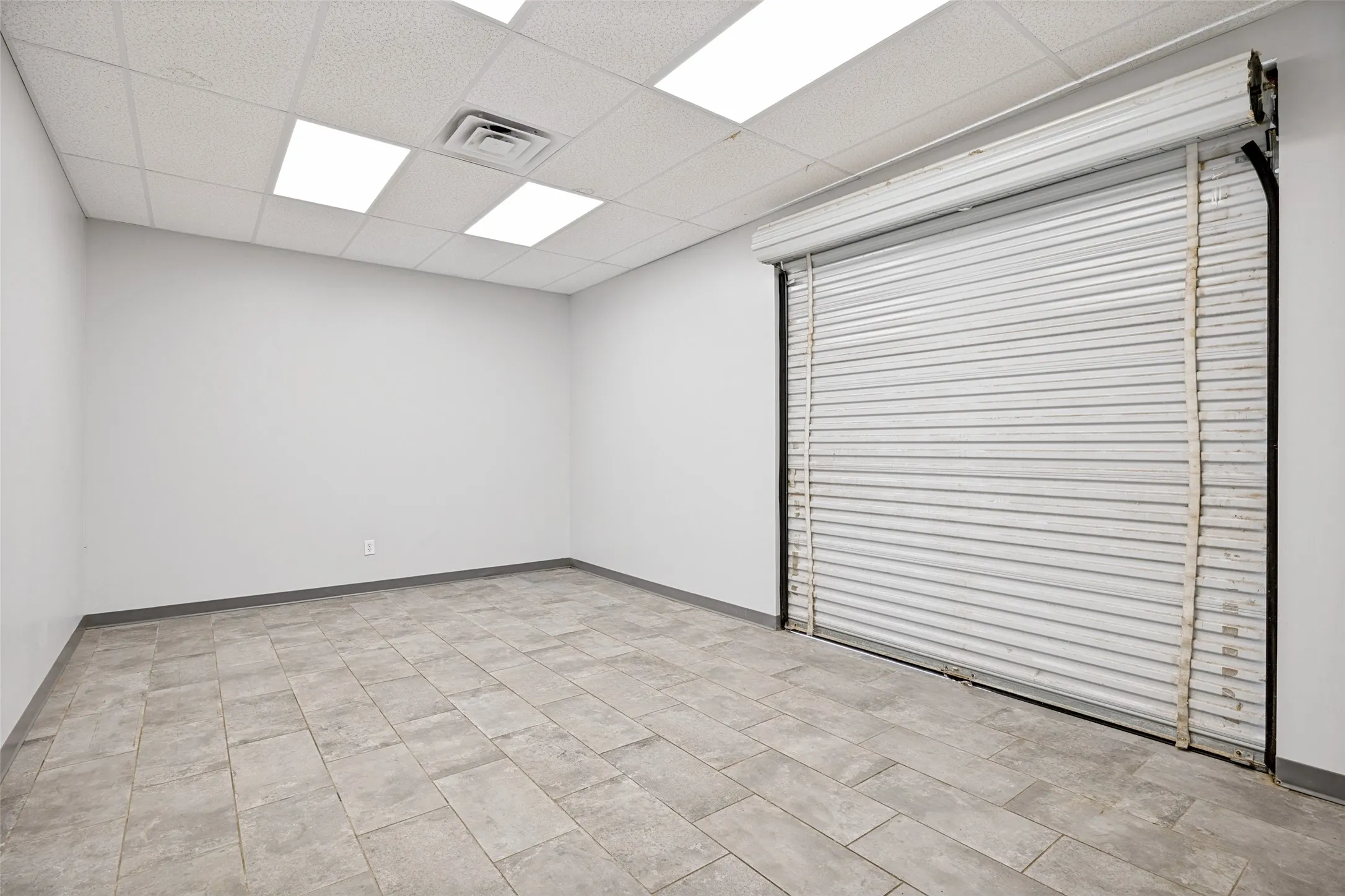 Storage/ supply room featuring a roll-up security door, tiled flooring, drop ceiling with recessed lighting.