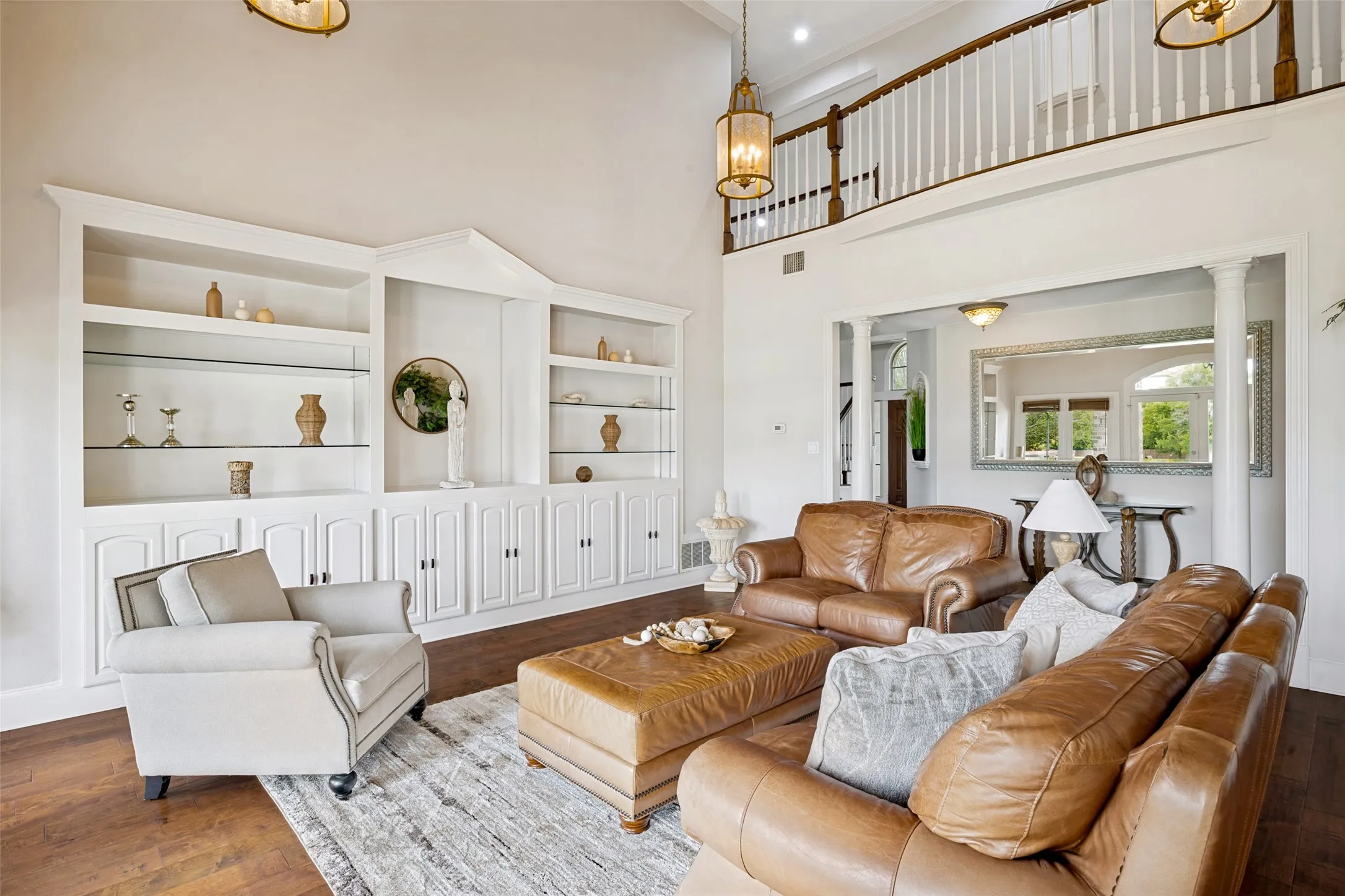 Living area featuring a two-story ceiling, built-in shelving with glass inserts, wood-finish flooring, a second-story balcony with white balusters, and a columned entryway