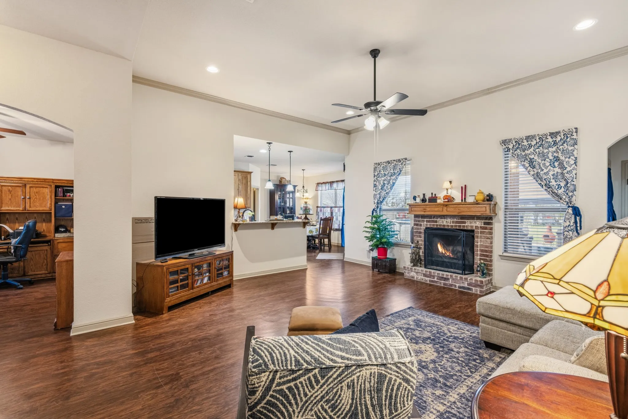 Spacious living area with warm wood flooring, crown molding, and a cozy brick fireplace. Open layout flows seamlessly into the kitchen and dining space, perfect for everyday living and entertaining.