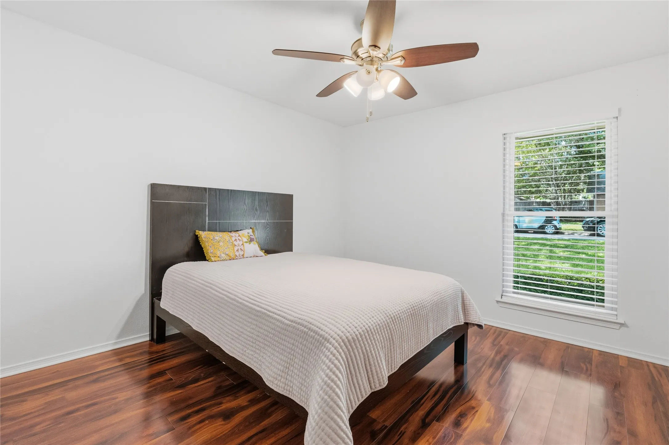 Bright interior space featuring rich wood-finish flooring, a ceiling fan with integrated lighting, and a single window with blinds