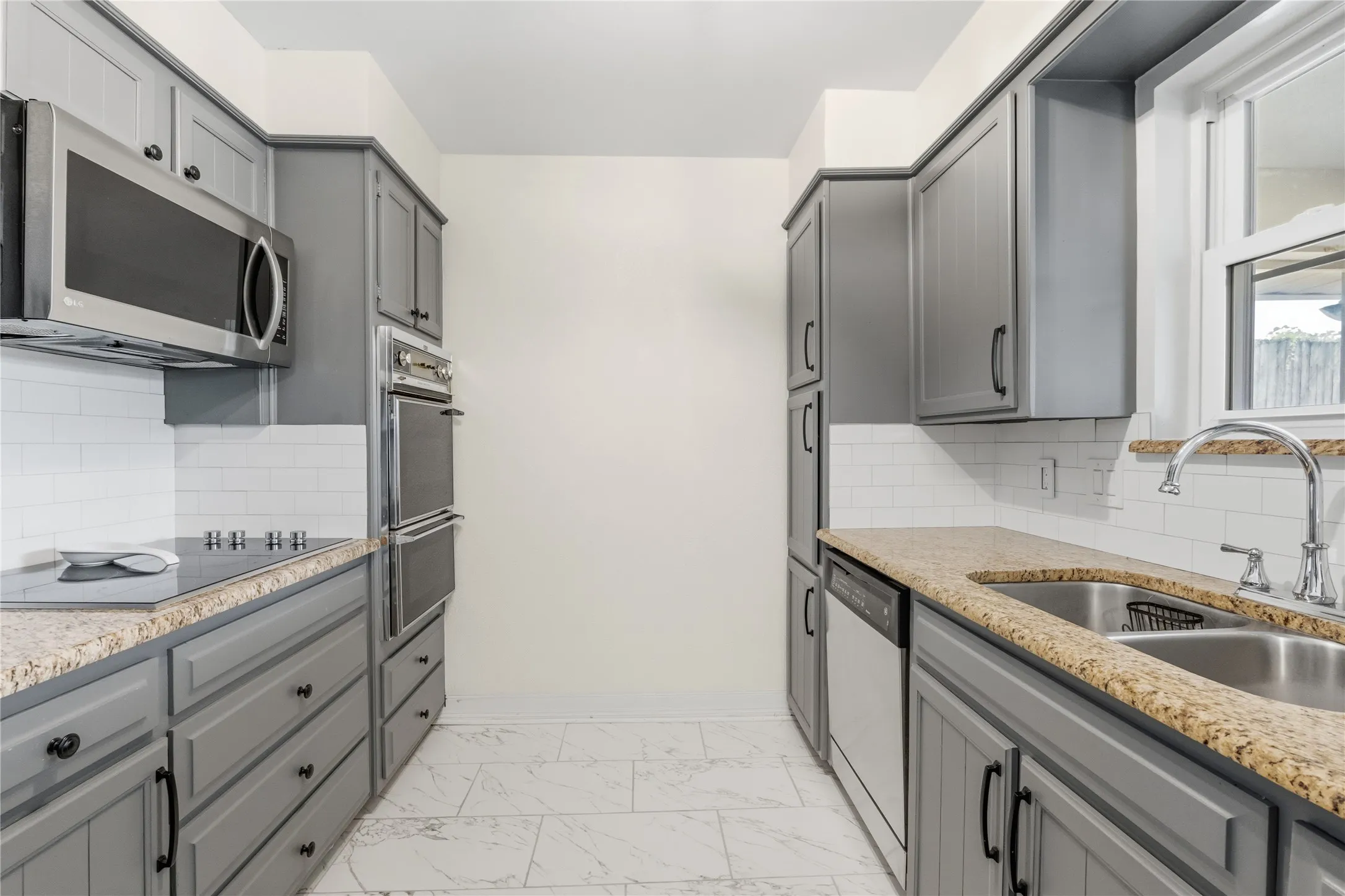Galley kitchen featuring gray cabinetry, stainless steel appliances, granite countertops, subway tile backsplash, and light-toned tile flooring