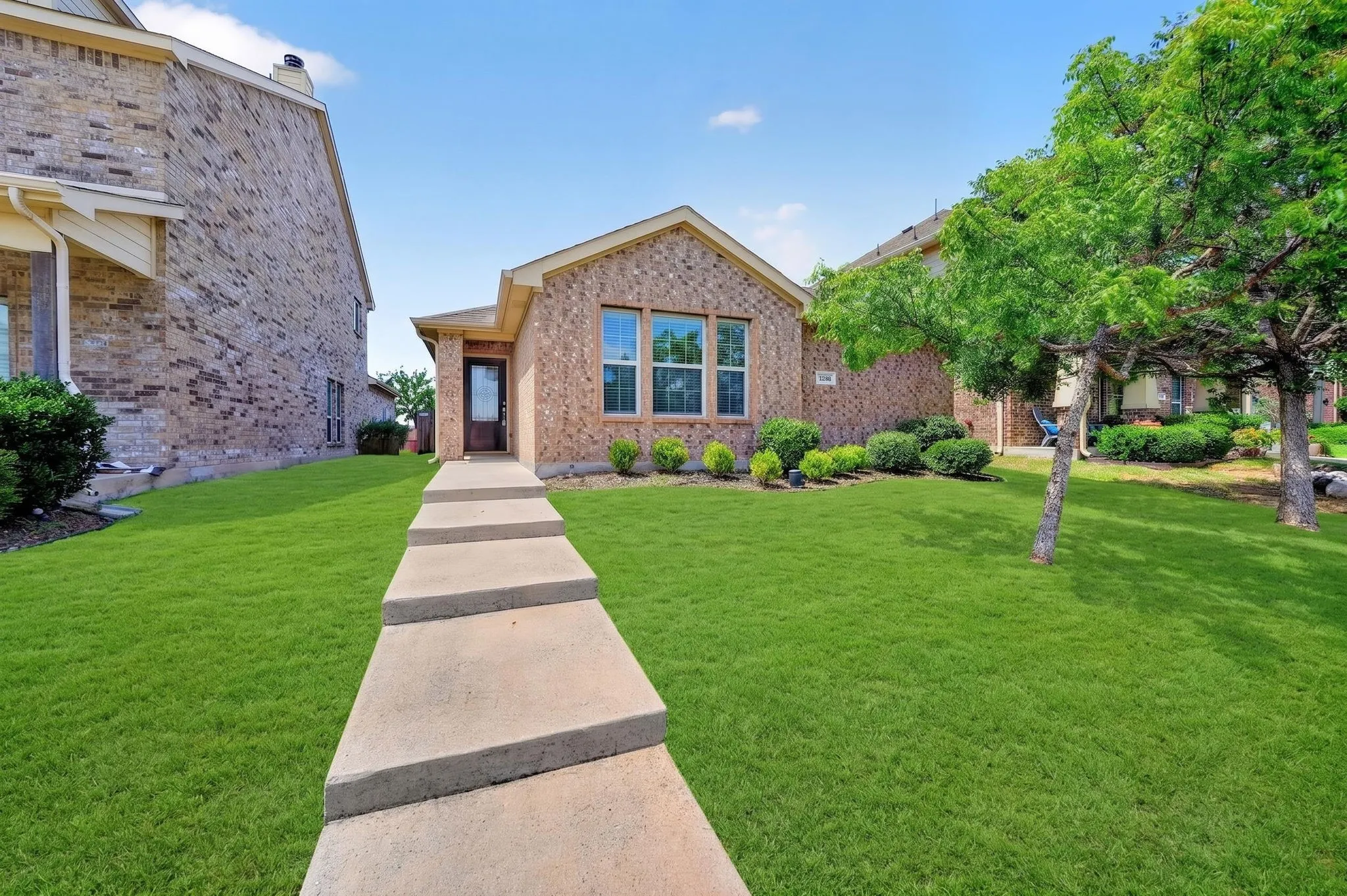 Brick exterior with a gabled roofline, a stepped concrete walkway, lush green lawn, and mature trees