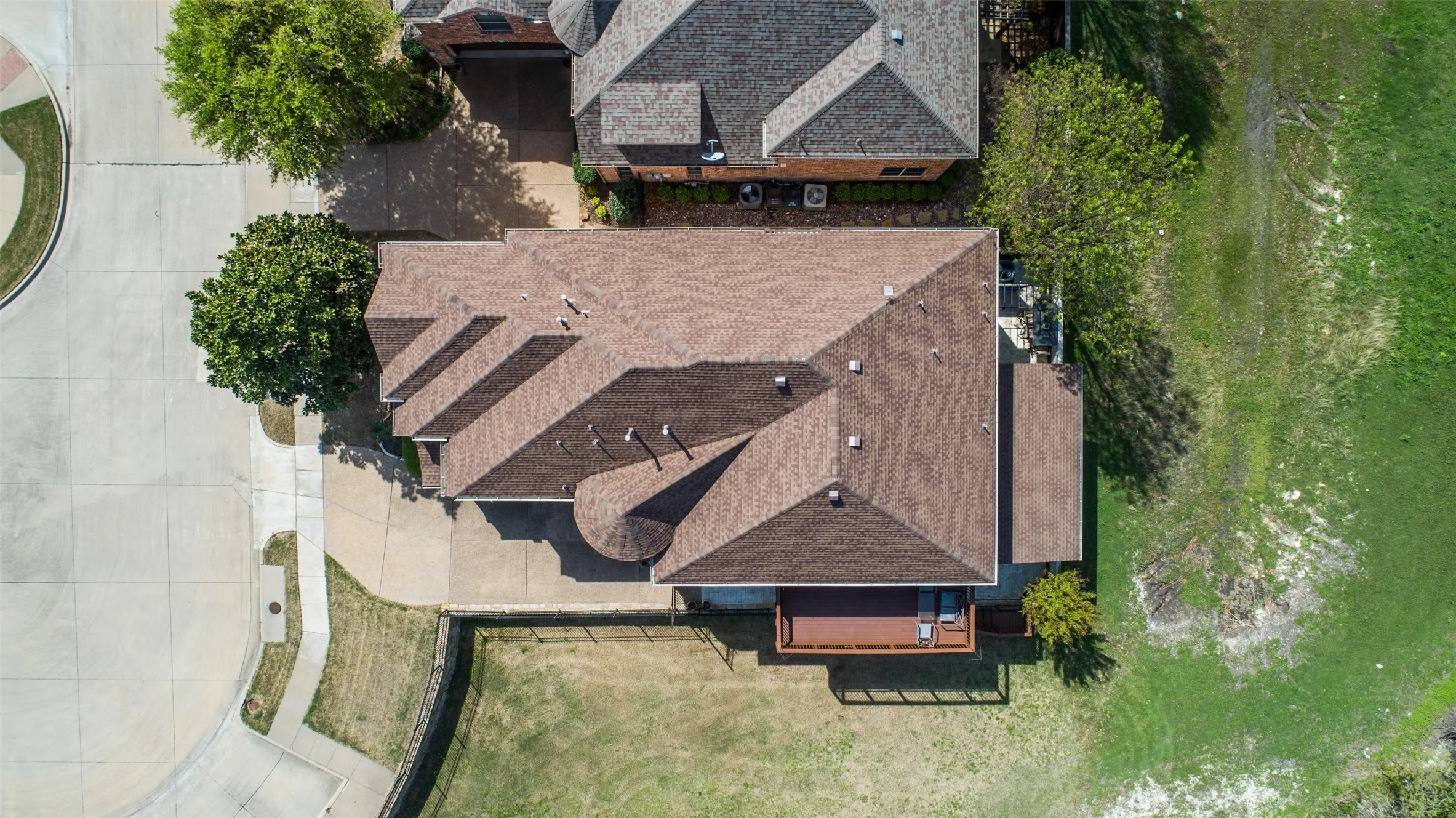 Aerial perspective showcasing a property with a brown shingle roof, an expansive concrete driveway, and a wood-finish deck
