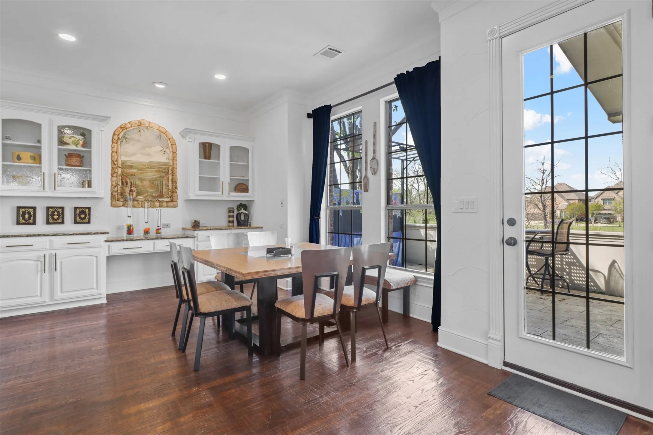 Dining area featuring rich wood-finish flooring, white built-in cabinetry with glass-front uppers, a built-in countertop desk, and a grid-paneled glass door leading to an outdoor patio