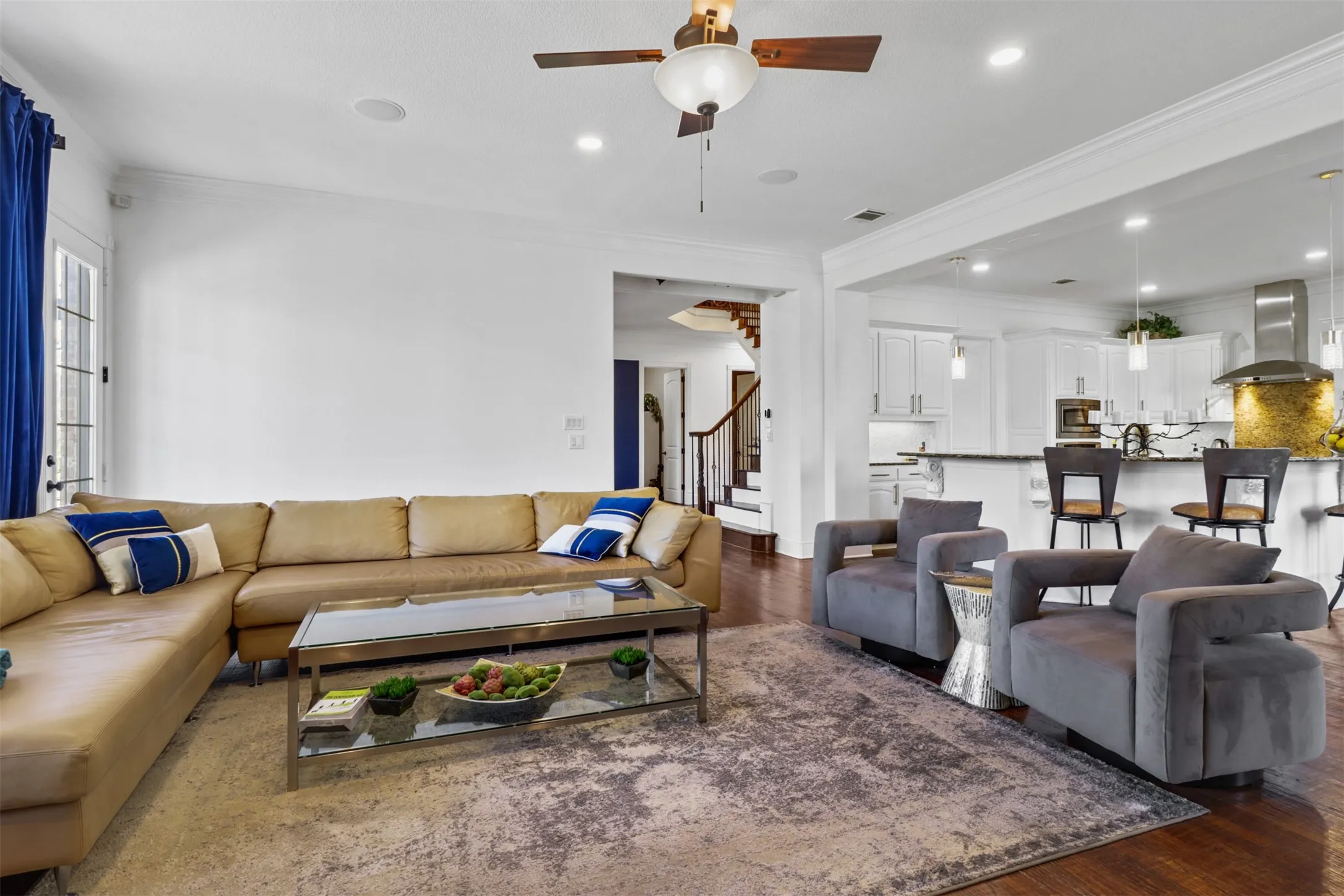 Open-concept living area featuring rich wood-finish flooring, recessed lighting, crown molding, a ceiling fan, and an expansive kitchen with white cabinetry