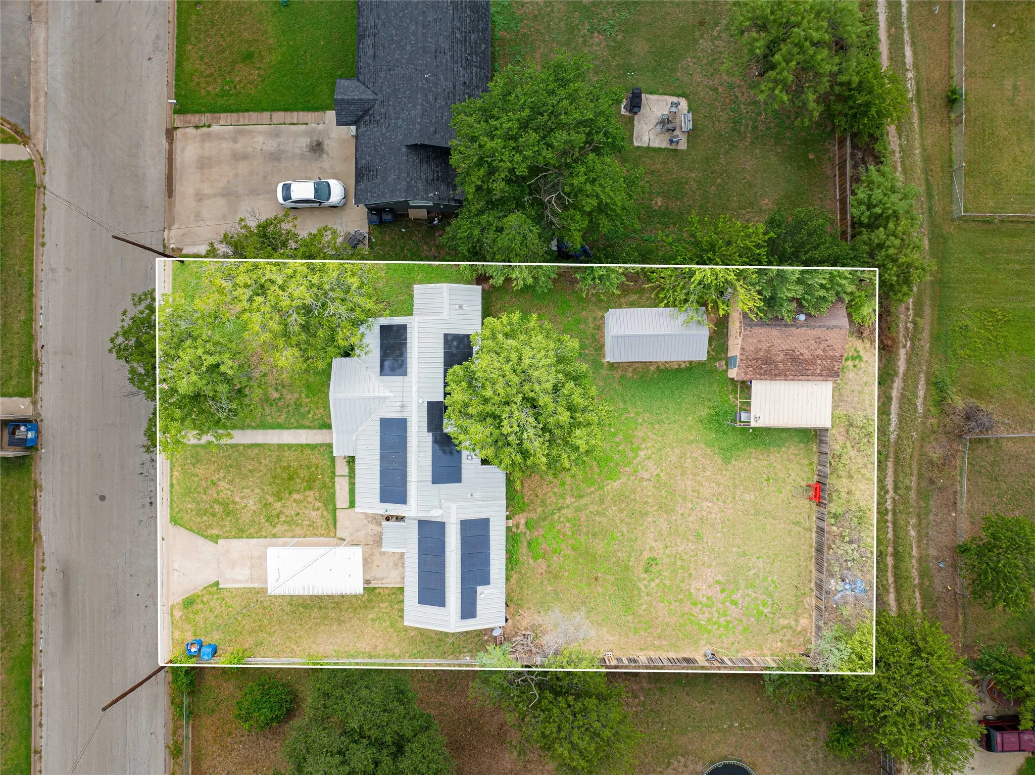 Overhead property view featuring a residence with multiple solar panels on the roof, a front yard with a concrete walkway, a gravel driveway, and mature trees