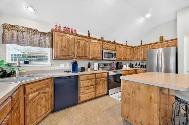 Kitchen featuring wood cabinetry, stainless steel appliances, a corner sink with window, an island, and tile flooring