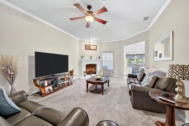 Living area featuring a fireplace with a white mantel, neutral wall paint, crown molding, and a ceiling fan