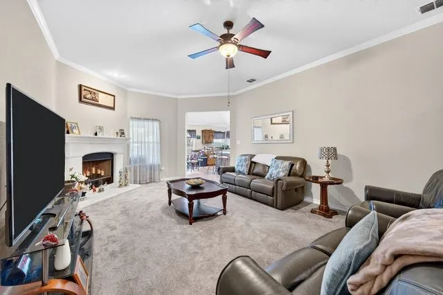 Living area featuring a fireplace with a light-colored mantel, crown molding, a ceiling fan with integrated lighting, and carpet flooring