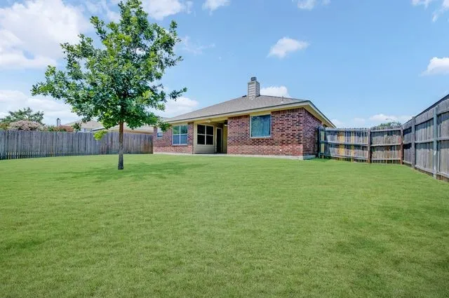 Expansive green lawn with mature tree, bordered by a wooden privacy fence, featuring a brick exterior home with a covered patio