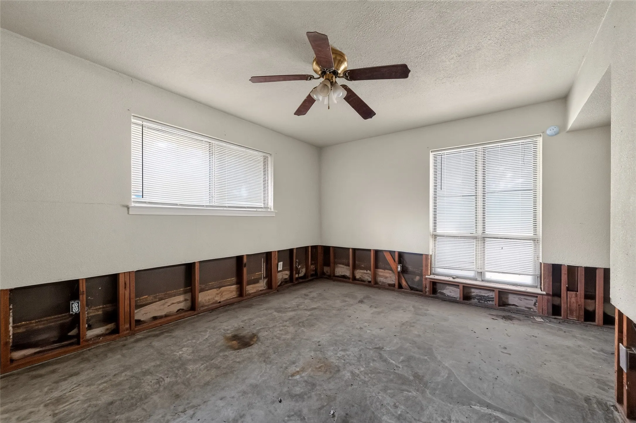 Room featuring two windows with horizontal blinds, a ceiling fan with light fixture, exposed wall studs, and concrete flooring