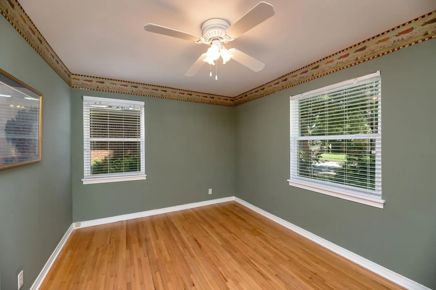 Hardwood flooring throughout, complemented by two windows with white blinds, a ceiling fan with light fixture, and crown molding