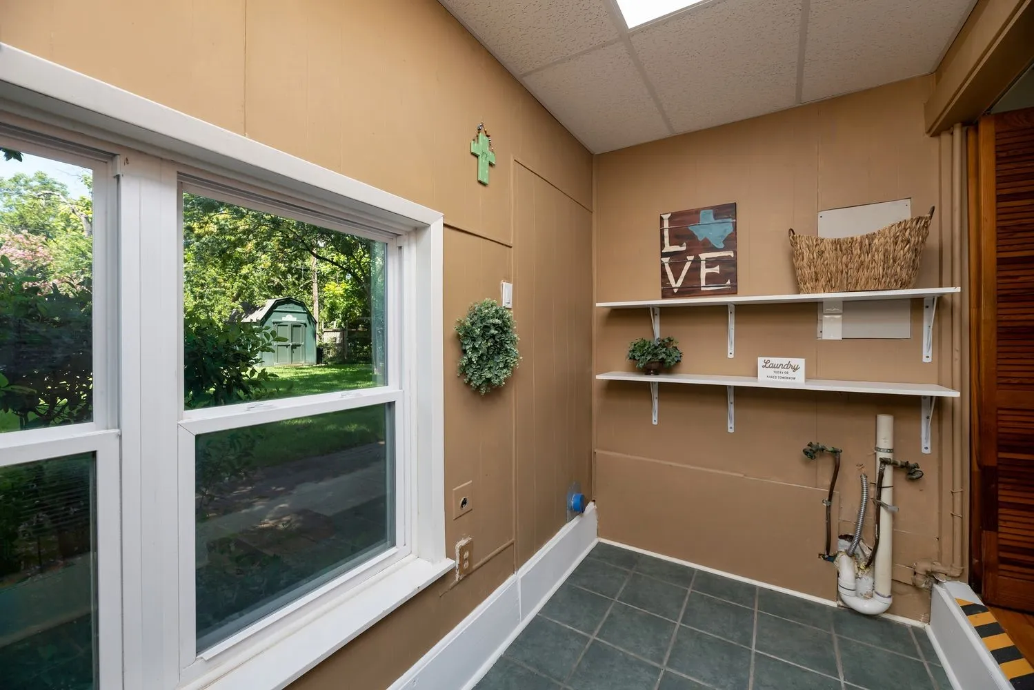 Utility space featuring a window overlooking green landscaping, tiled flooring, built-in shelving, and exposed plumbing hookups
