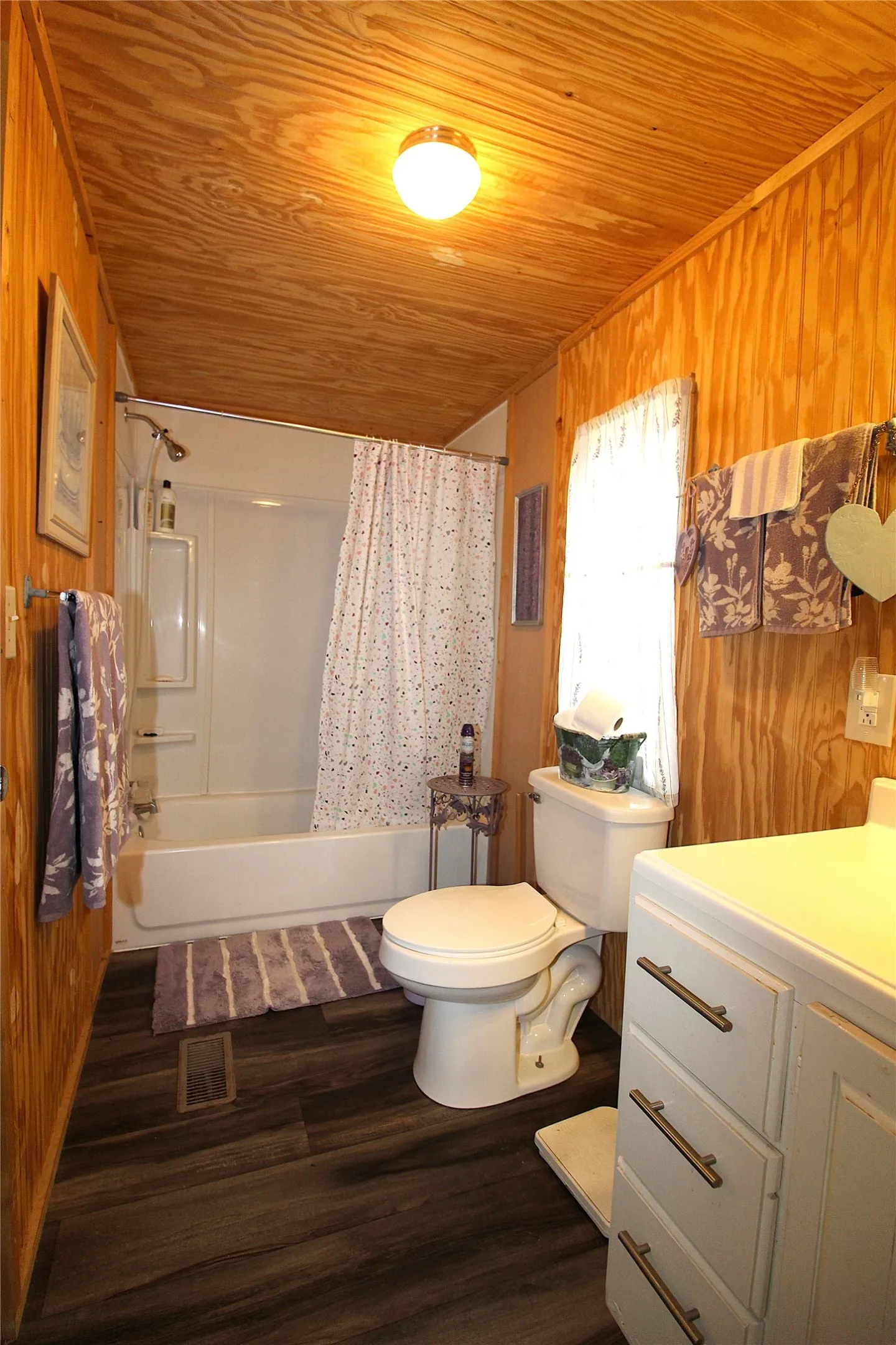 Bathroom featuring wood-paneled walls and ceiling, dark wood-finish flooring, a white bathtub with shower, a white toilet, and a white vanity with drawers
