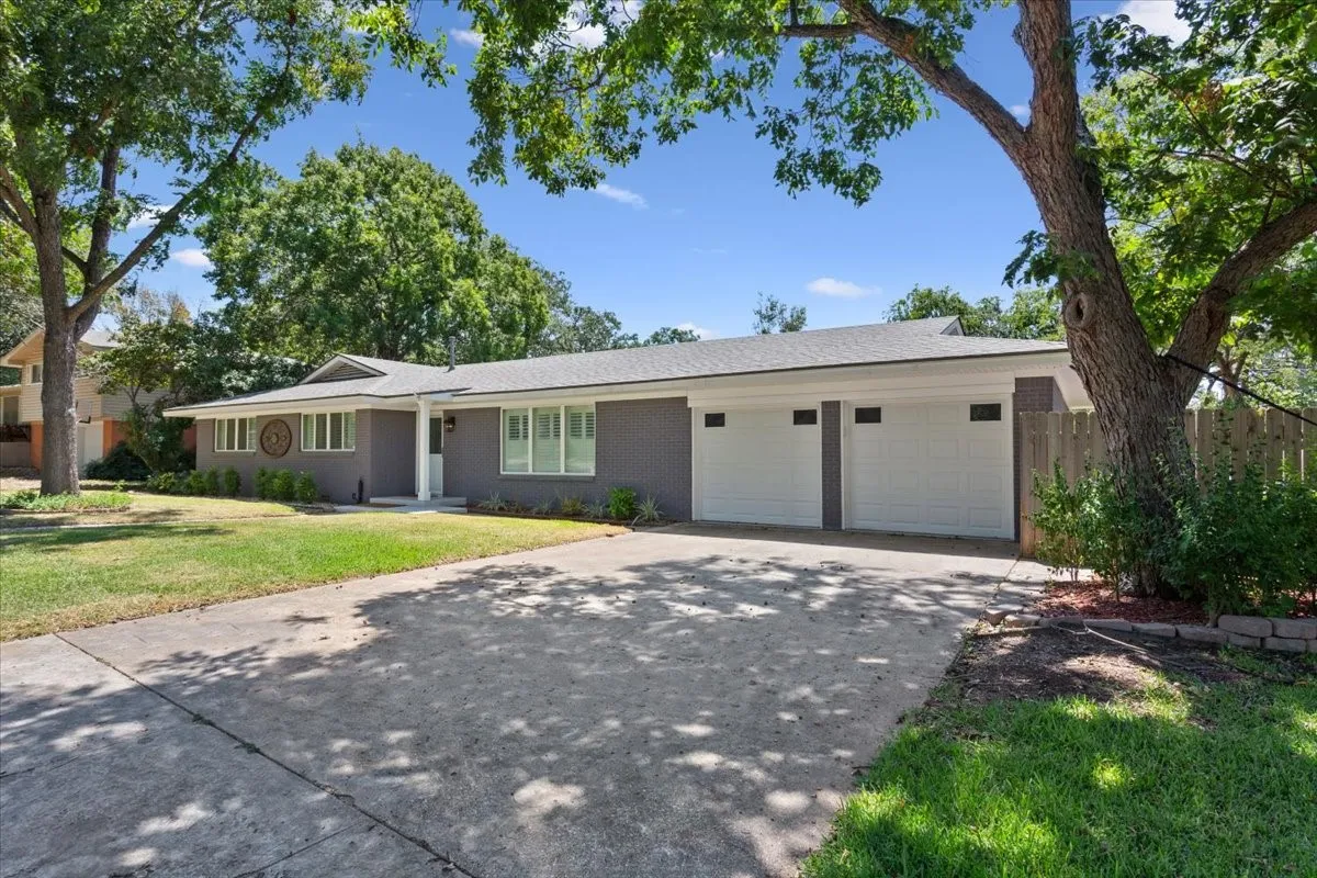 Gray brick exterior with white trim, two white garage doors, a concrete driveway, and a front lawn with mature trees
