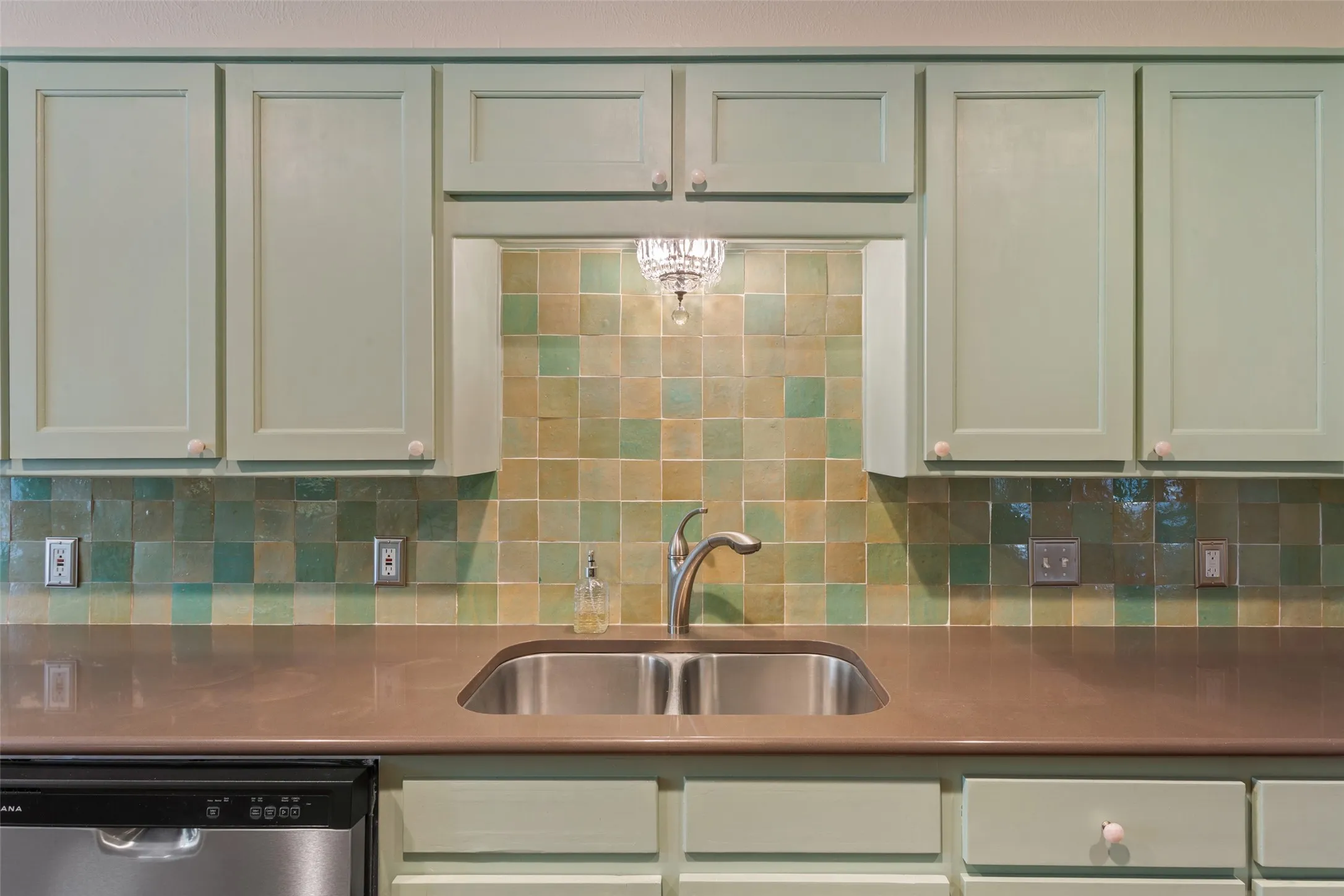 Updated kitchen featuring cream-colored cabinetry with recessed panel doors, stainless steel undermount sink, solid surface countertop, and a multi-colored ceramic tile backsplash in warm earth tones