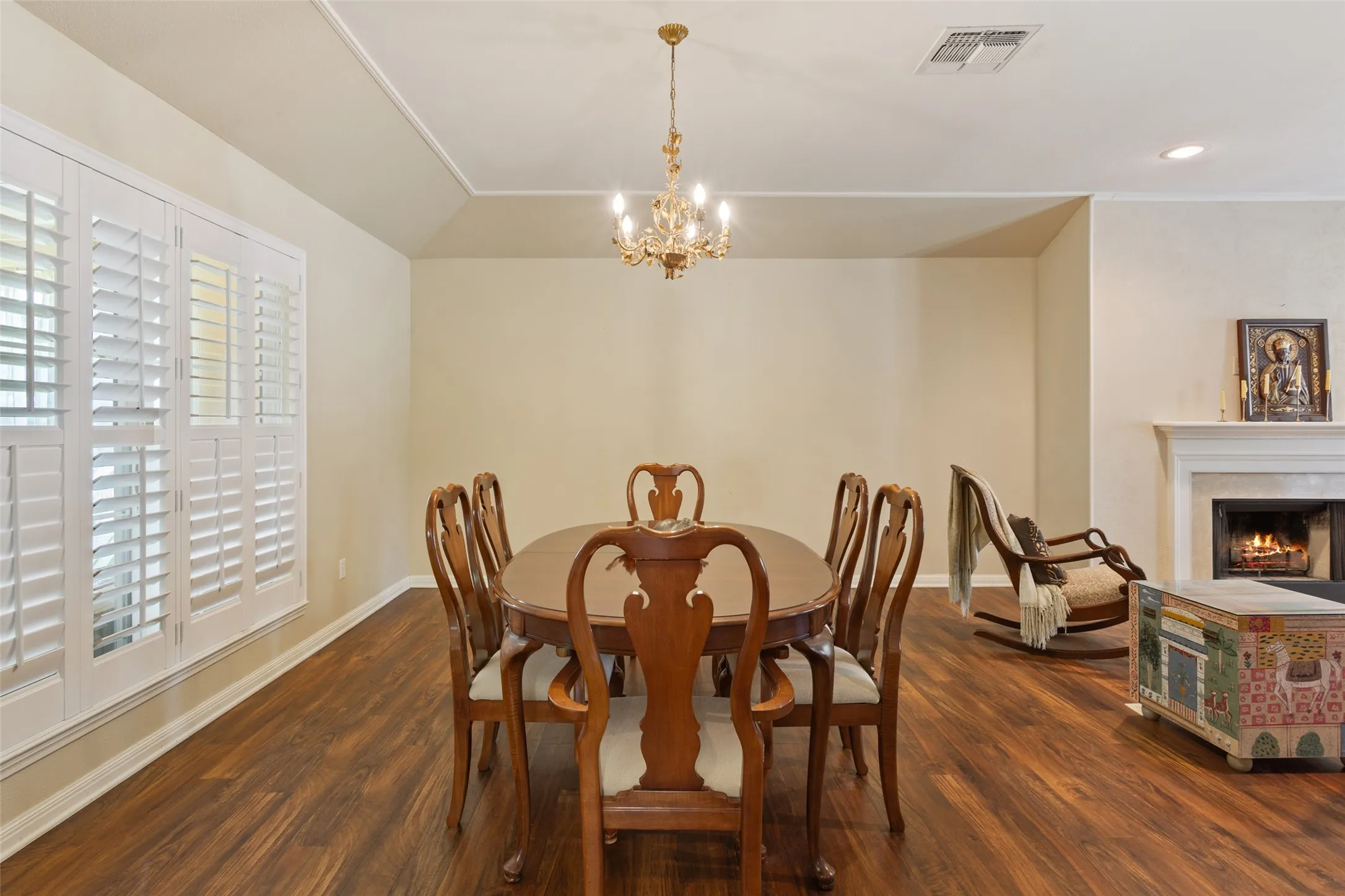 Wood-finish flooring with a fireplace featuring a white mantel, ceiling chandelier, and plantation shutters