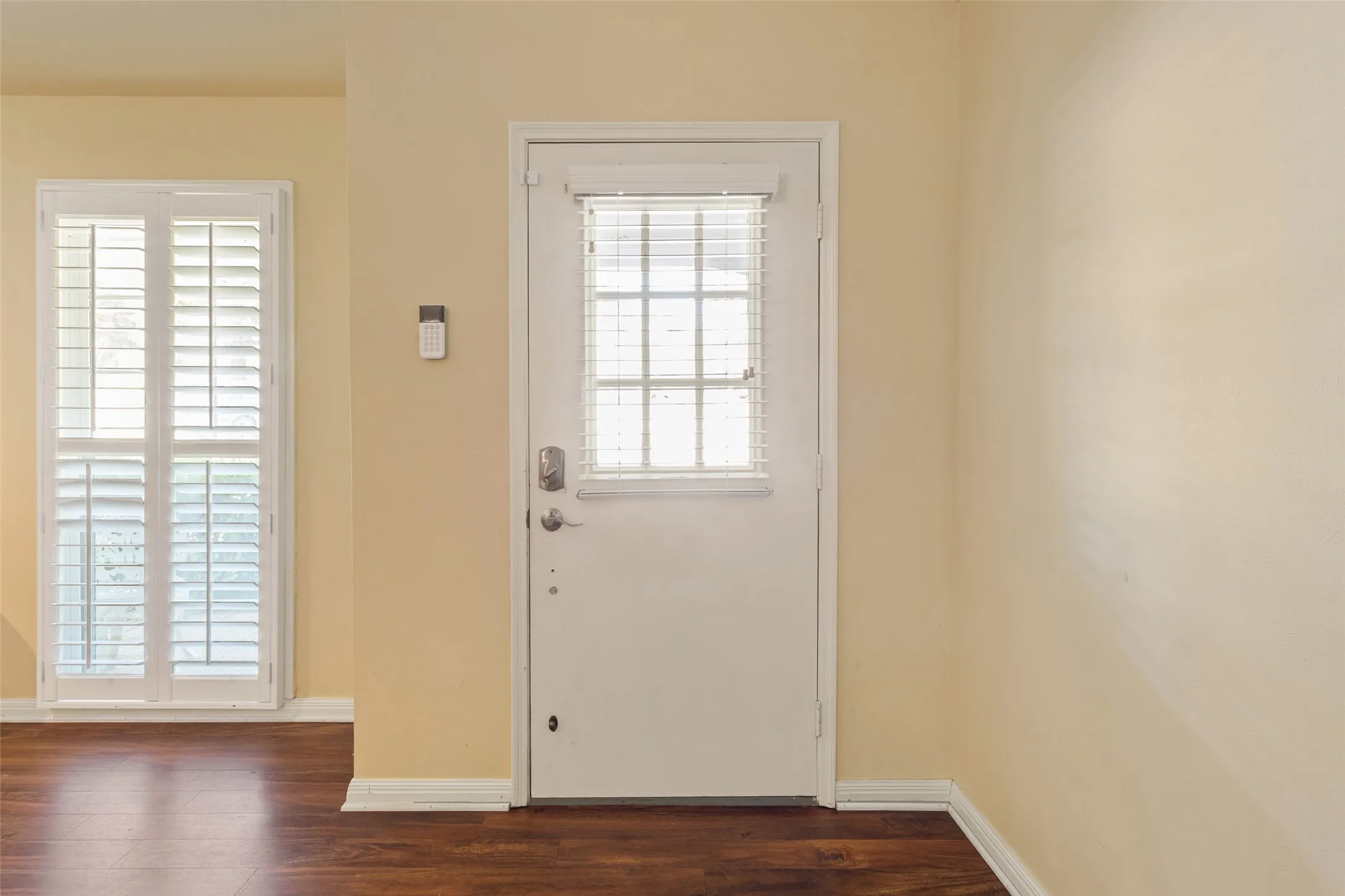 Wood-finish flooring with baseboard trim, and a white door featuring a window with blinds