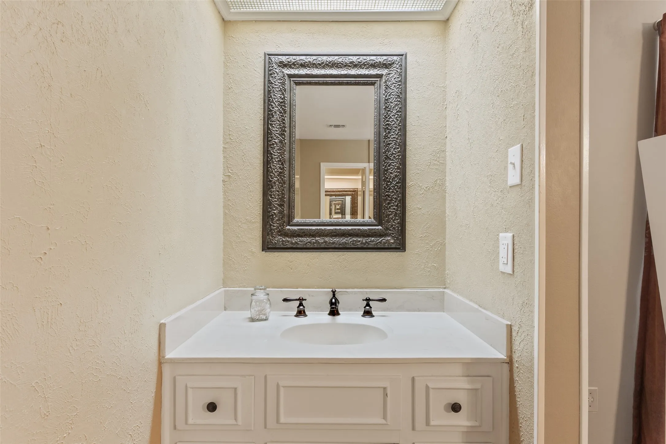 Elegant powder room featuring a white porcelain sink with oil-rubbed bronze faucet and hardware, cream-colored cabinetry with recessed panel doors, textured cream walls, and an ornate silver-framed mirror reflecting soft overhead lighting