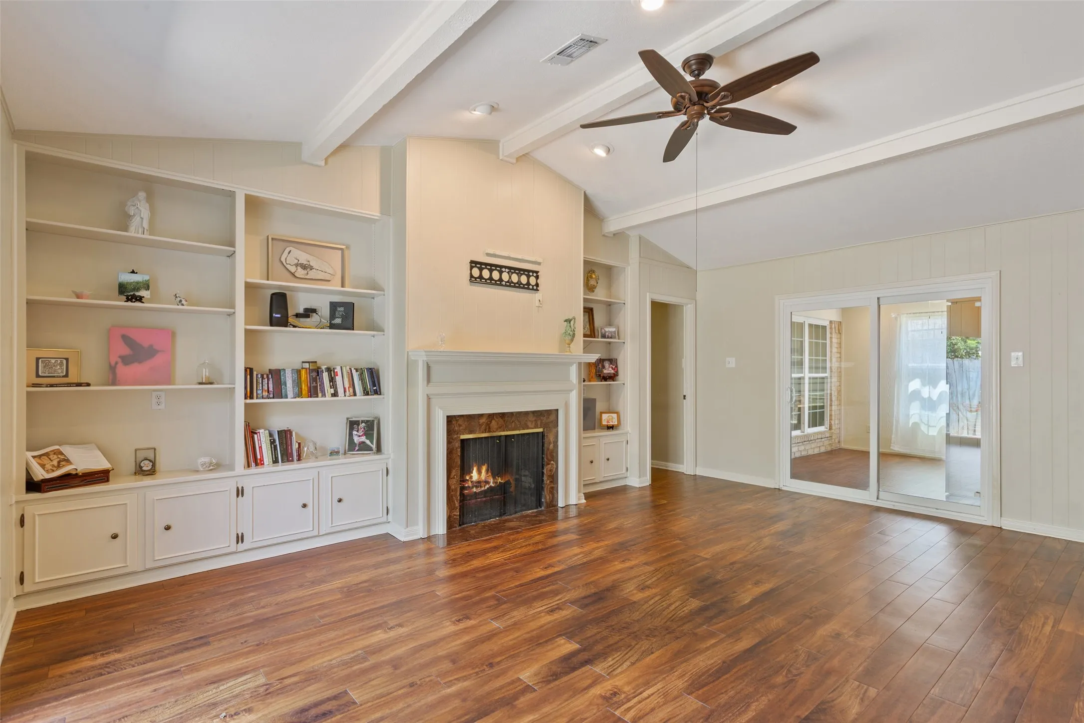 Vaulted ceiling with exposed beams, wood-finish flooring, built-in shelving and cabinetry, fireplace with mantel, and sliding glass doors