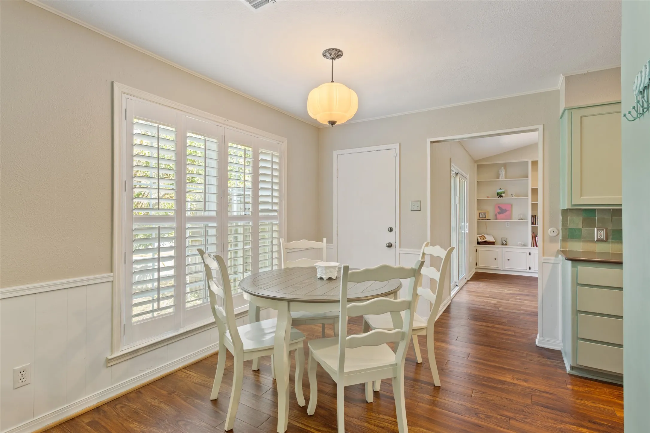 Wood-finish flooring, white plantation shutters, white wainscoting, and a built-in bookshelf with lower cabinetry