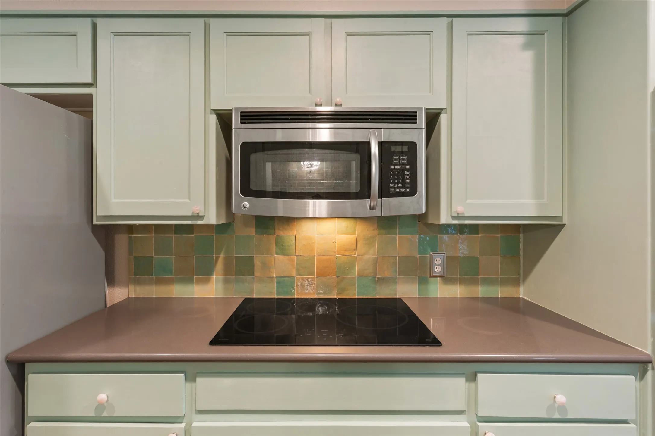 Updated kitchen featuring soft green cabinetry with recessed panel doors, stainless steel over-the-range microwave, solid surface countertops, and a geometric tile backsplash in earth tones with under-cabinet lighting