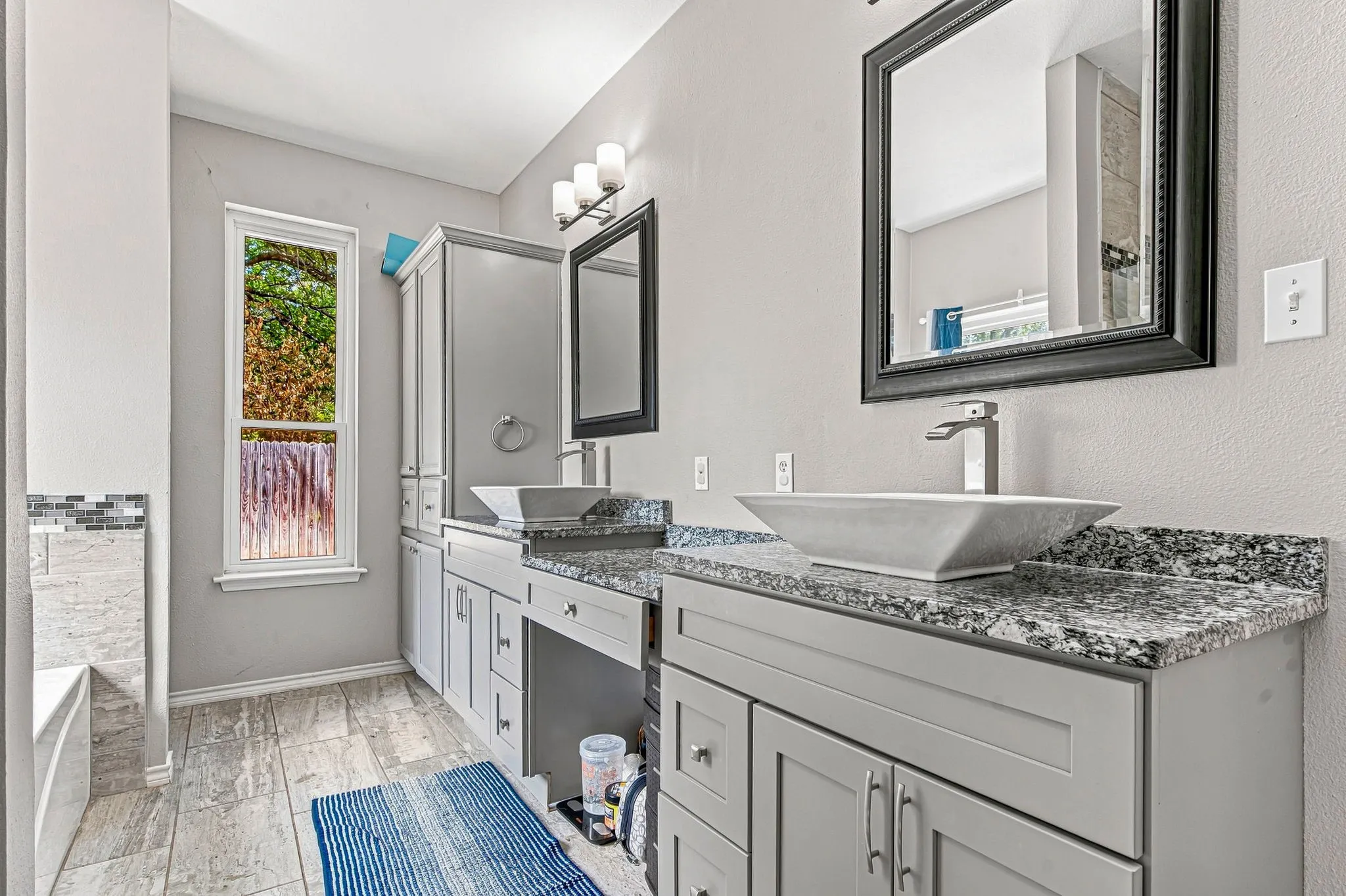 Dual vessel sinks with modern faucets, framed mirrors, stone countertops, light gray cabinetry, and tile flooring