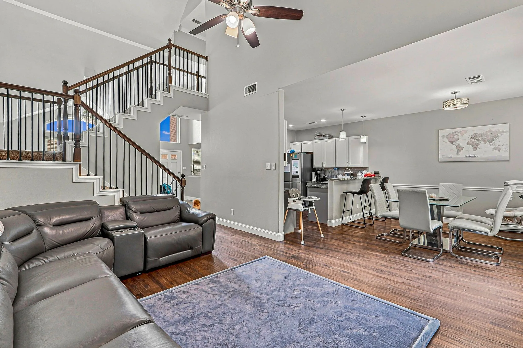 Vaulted ceiling with a ceiling fan, wood-finish flooring, two-story staircase with dark wood handrails and black metal balusters, recessed lighting, and kitchen with white cabinetry and pendant lighting