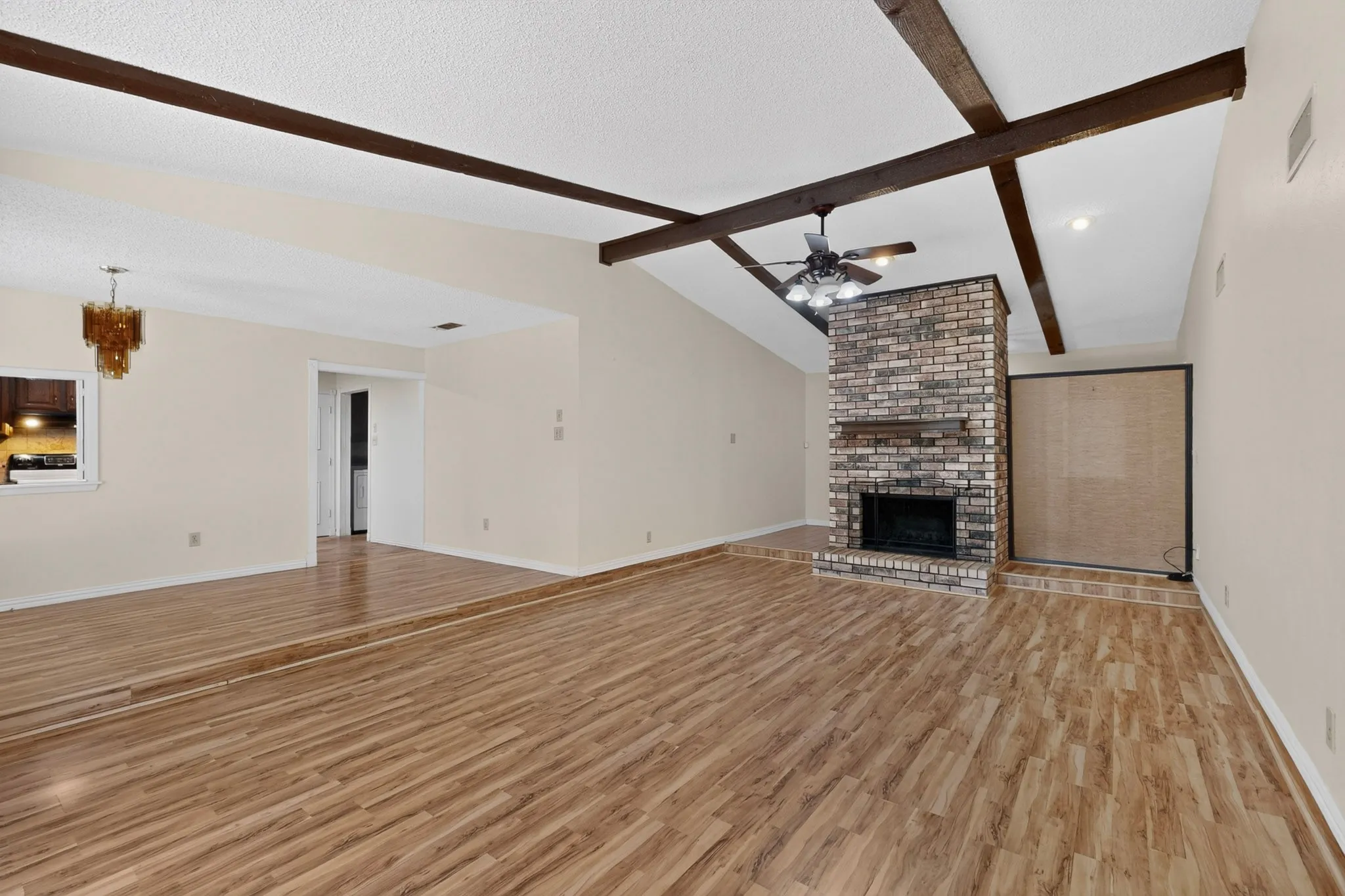 Wood-finish flooring throughout, vaulted ceiling with exposed beams, brick fireplace with mantel, and a ceiling fan with integrated lighting