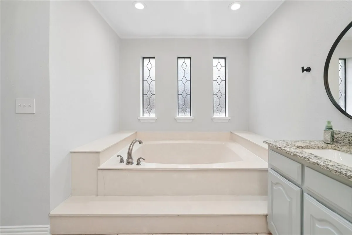Built-in soaking tub with tiered surround, three decorative leaded glass windows, recessed ceiling lighting, and a vanity with a granite countertop and undermount sink