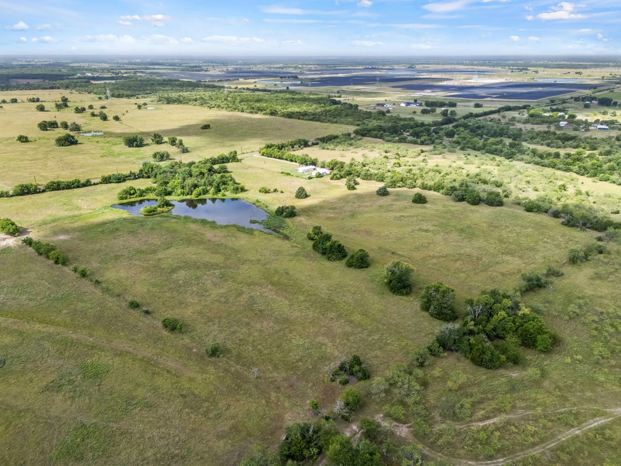 Expansive rural landscape featuring a natural pond, scattered trees, and open fields under a blue sky with clouds